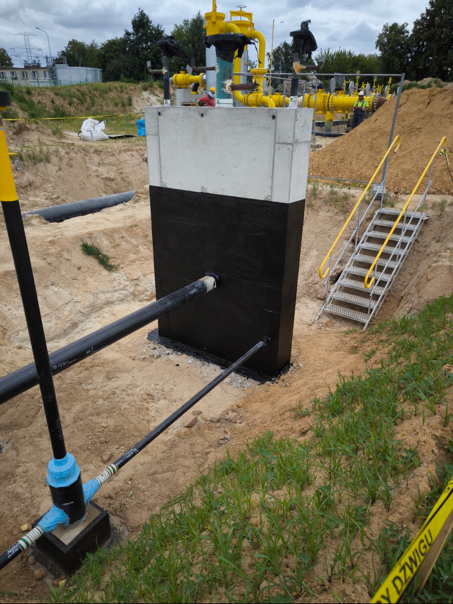 Betonowa konstrukcja z izolacją bitumiczną, rurami i instalacją gazową w wykopie, metalowe schody z żółtymi poręczami prowadzą w dół, widoczne taśmy ostrzegawcze.