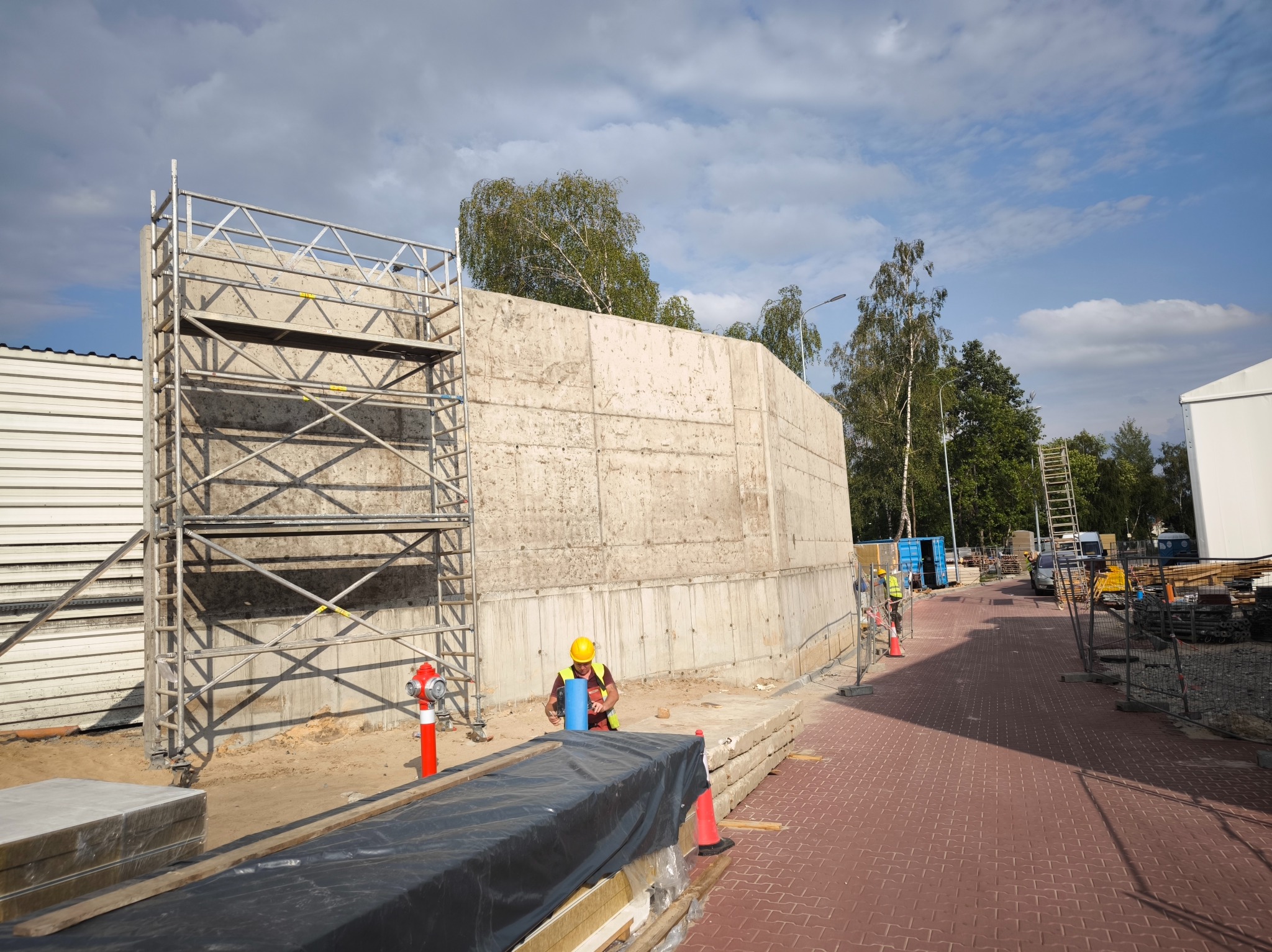 Pracownik w kasku i kamizelce odblaskowej pracuje na budowie przy betonowej ścianie, widoczne rusztowanie, hydrant i czerwona kostka brukowa.