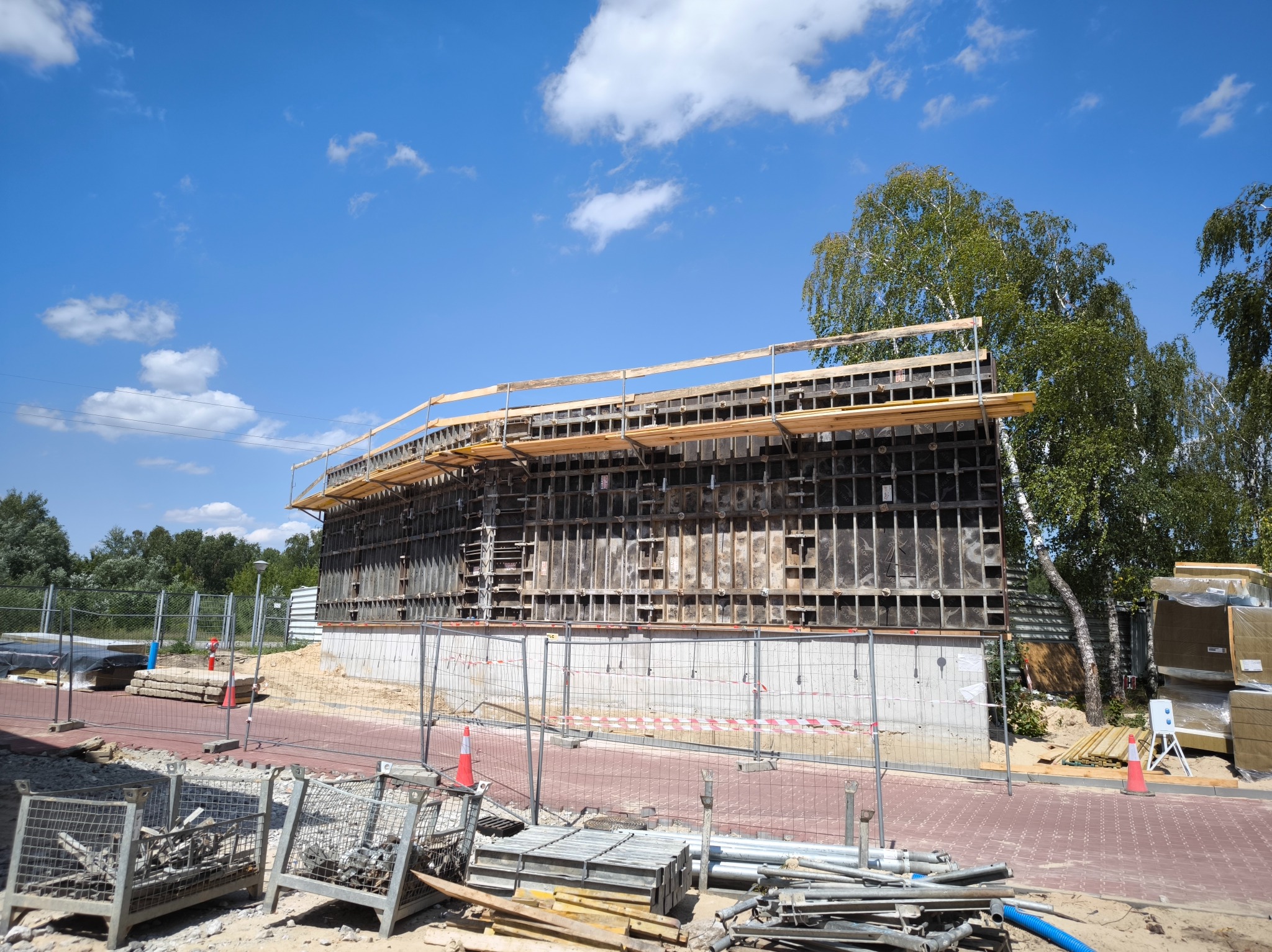 Szare szalunki pionowe z drewnianym rusztowaniem na tle błękitnego nieba z chmurami, obok drzewa i materiały budowlane, zabezpieczone siatką.