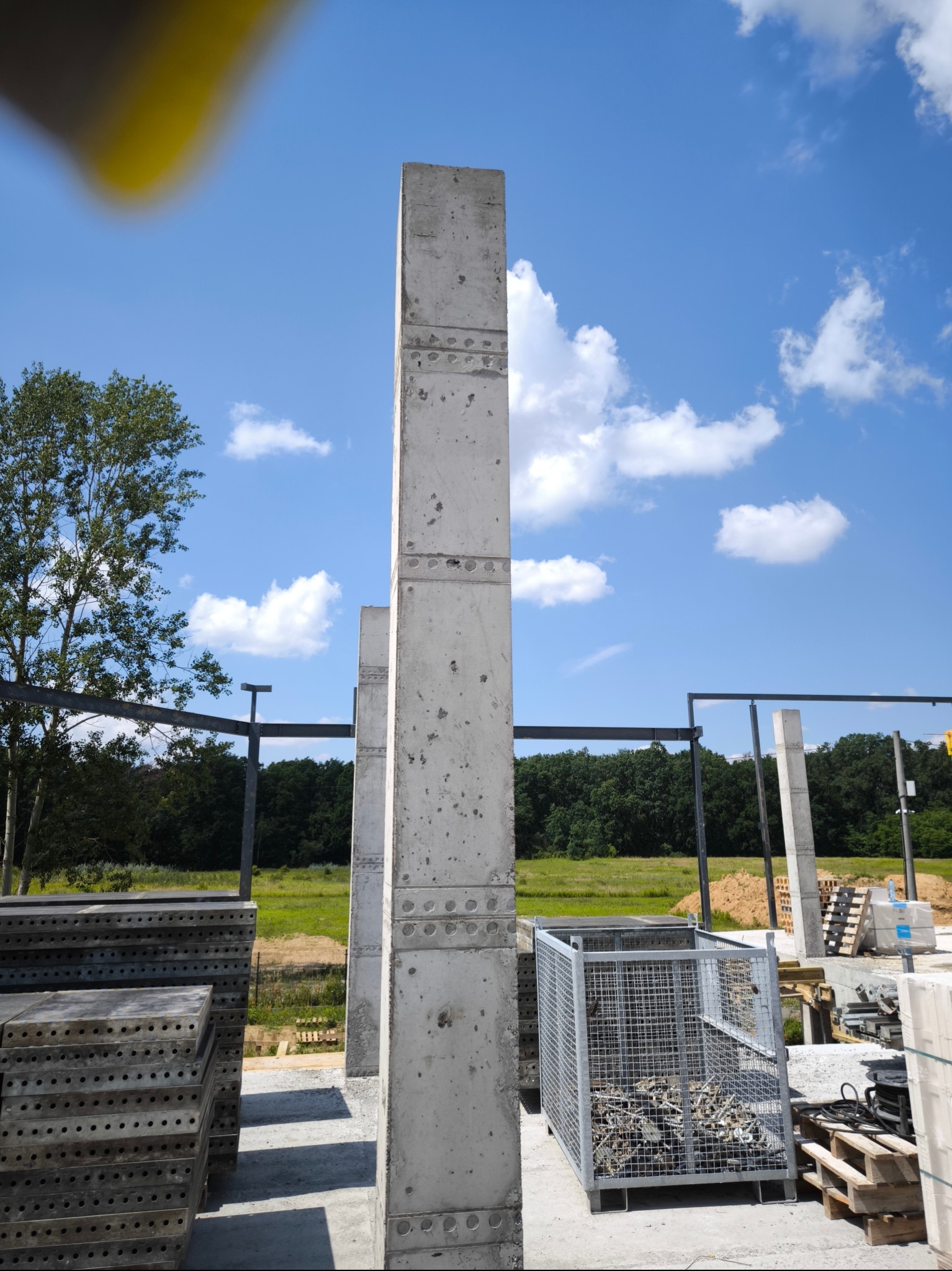 Wzniesione betonowe słupy konstrukcyjne na placu budowy, widoczne szalunki i materiały budowlane, w tle błękitne niebo z chmurami i zieleń drzew.