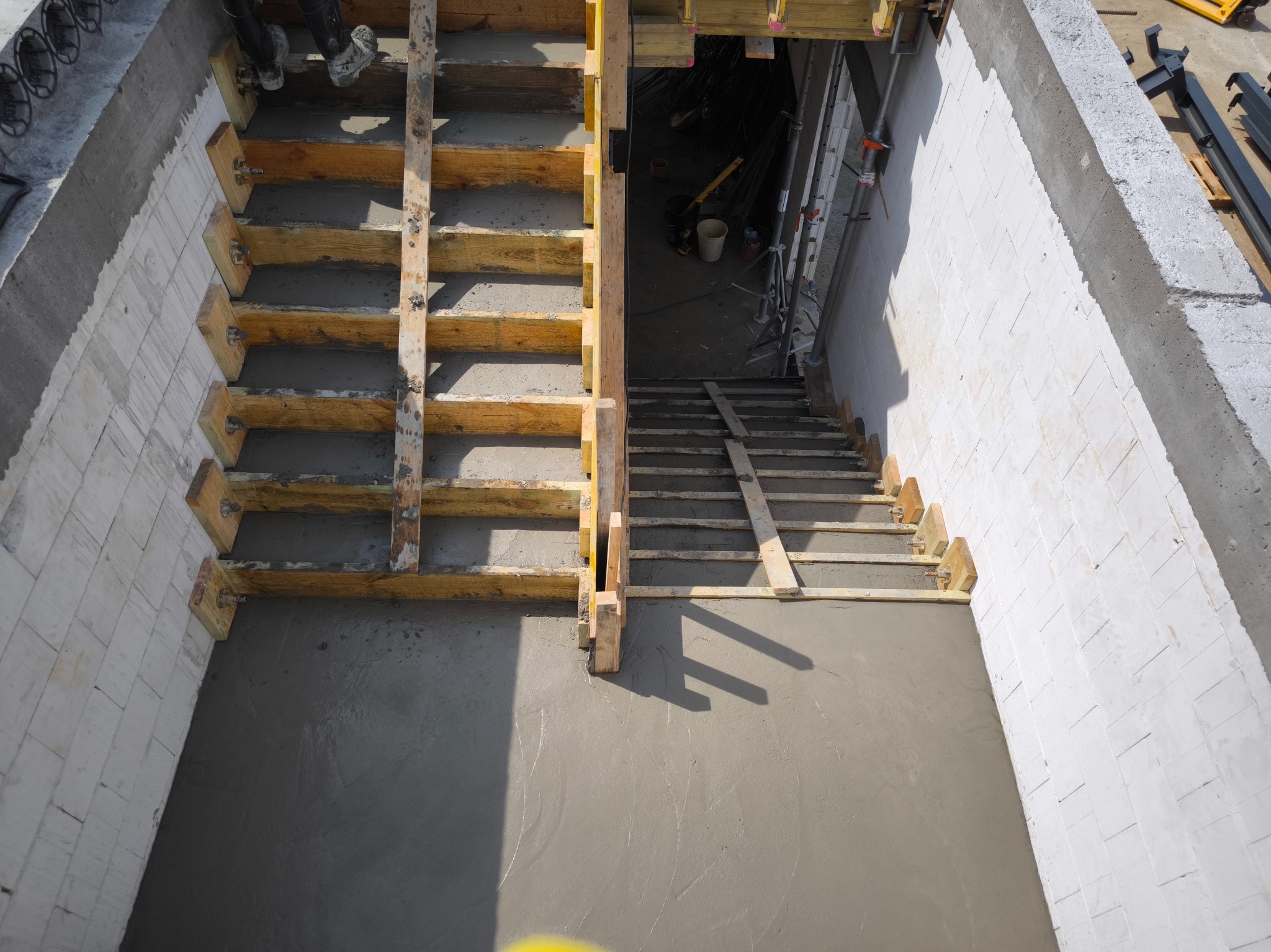 Wylana betonowa podłoga z drewnianym szalunkiem schodów, widok z góry na klatkę schodową w surowym stanie, białe ściany z bloczków betonowych, etap budowy.