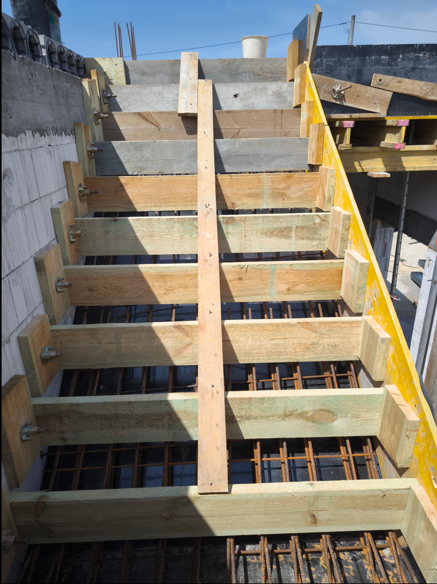 Szkielet schodów z desek i zbrojenia stalowego, widok z góry, konstrukcja szalunkowa z żółtymi elementami, przygotowanie do wylewki betonu.