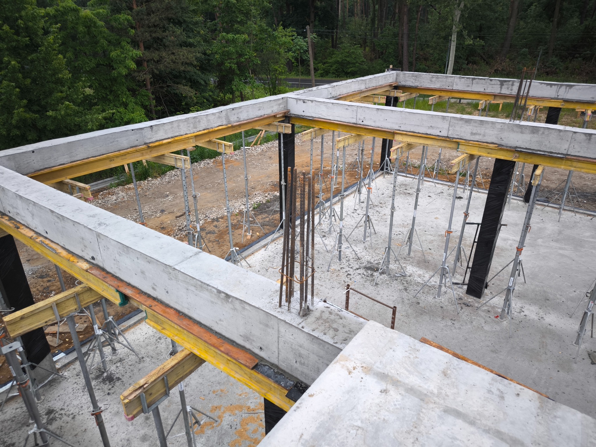 Wysoki kąt widzenia na szkielet budynku w trakcie budowy, z widocznymi stalowymi prętami zbrojeniowymi i systemem podpór szalunkowych, otoczony zielenią drzew.