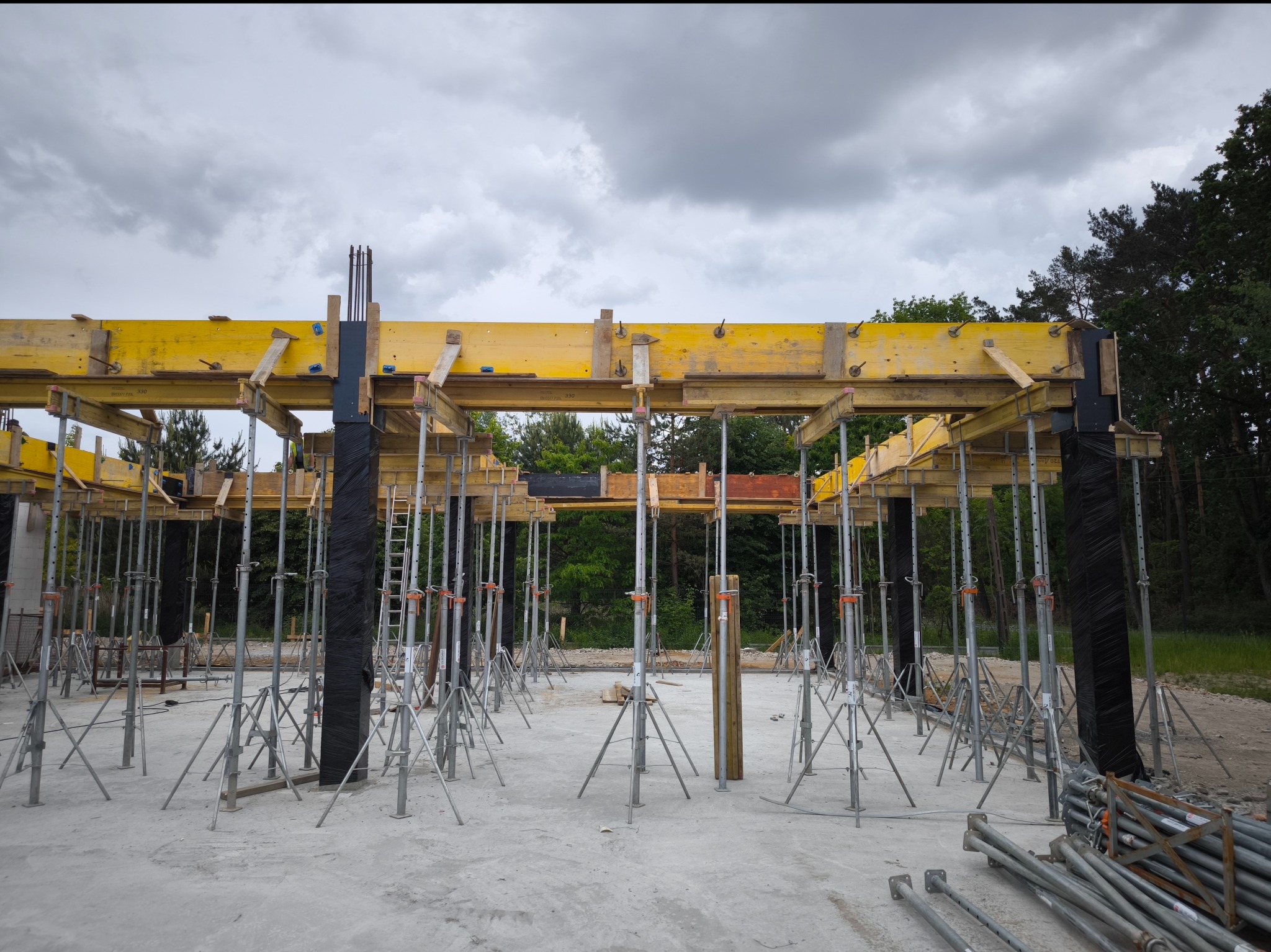 Szare niebo nad budową: Żółte szalunki wsparte na regulowanych podporach tworzą konstrukcję stropu, w tle widoczne drzewa.