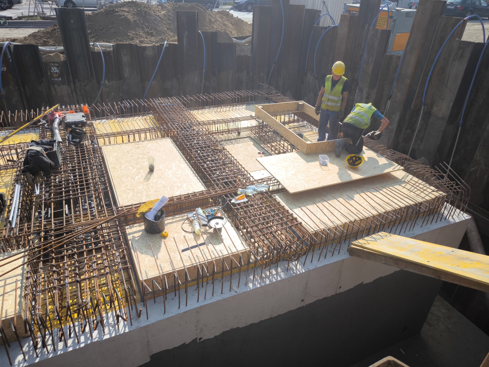 Budowa fundamentów: Widok z góry na wykop zabezpieczony ściankami Larsena, zbrojenie fundamentu z użyciem siatki stalowej i drewnianych szalunków, dwóch robotników w żółtych kamizelkach i kaskach...