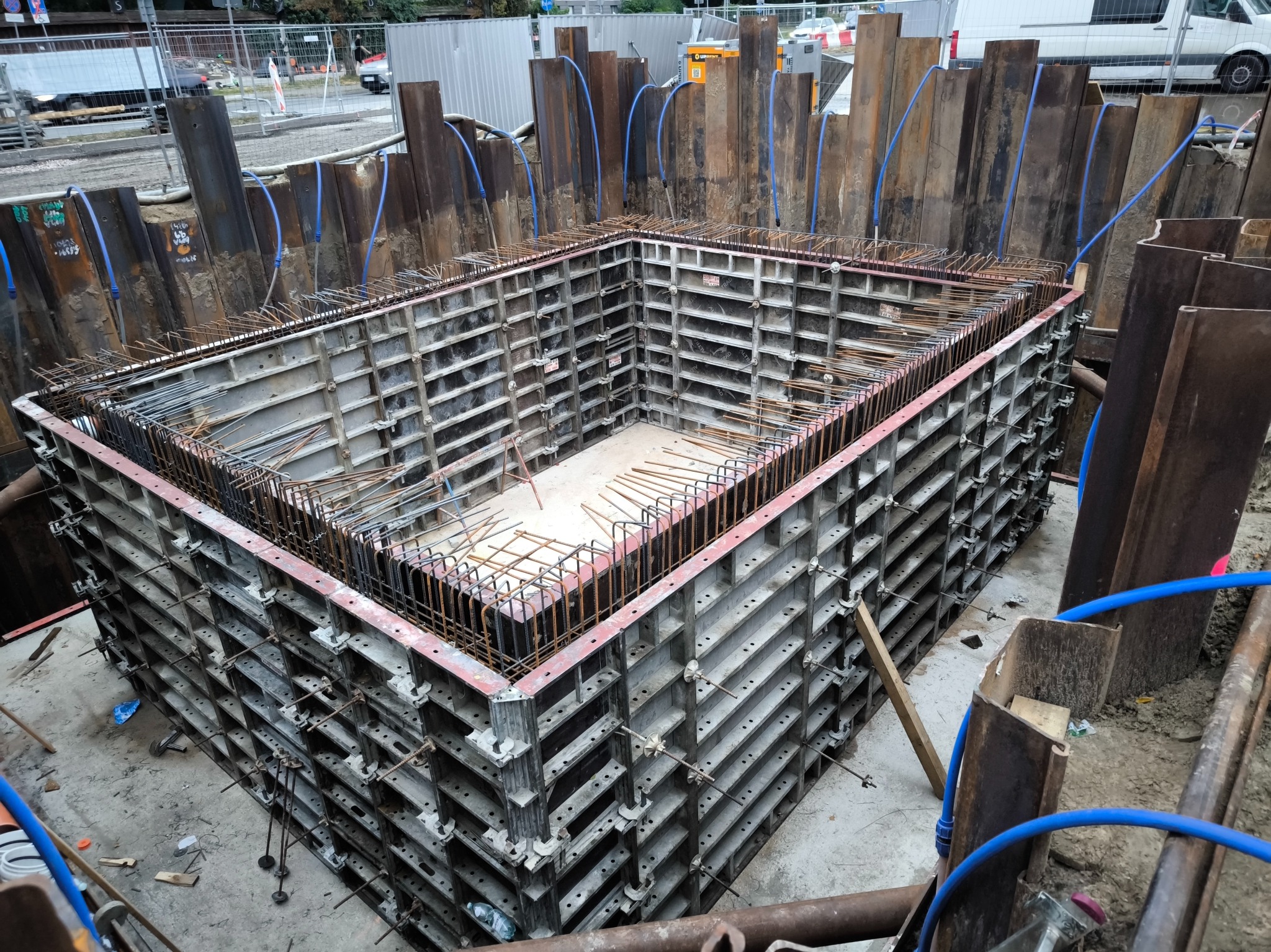 Szczegółowy widok szalunku fundamentowego z widocznym zbrojeniem w wykopie, zabezpieczonym stalowymi ściankami Larsena i niebieskimi przewodami odwadniającymi.