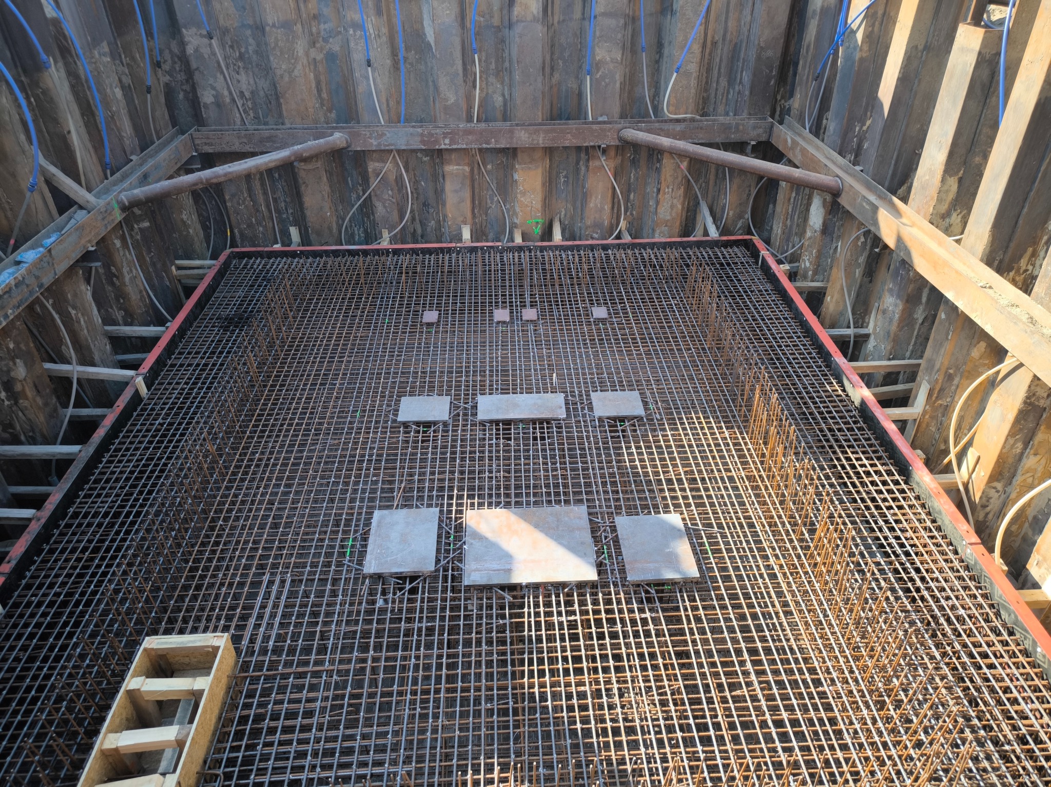 Siatka zbrojeniowa w wykopie, wzmocniona metalowymi płytkami, przygotowana do zalania betonem, z widocznym drewnianym szalunkiem i niebieskimi rurami w tle.