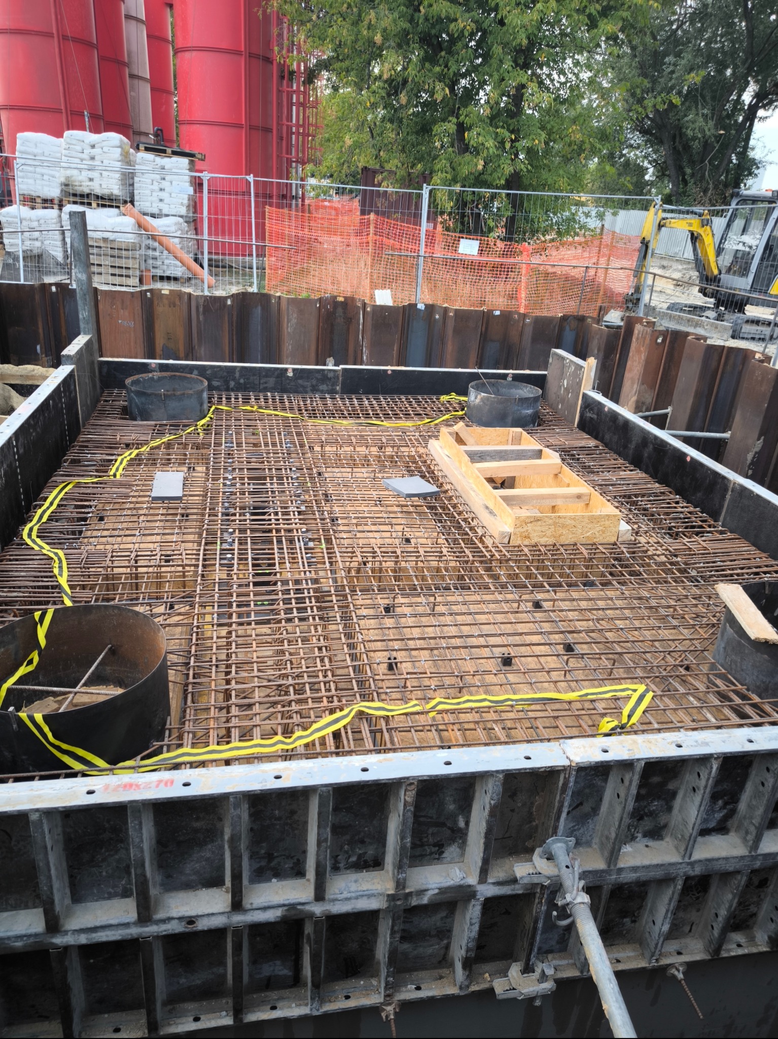 Zbrojenie fundamentu w wykopie z szalunkiem systemowym, widoczne elementy drewniane, żółta taśma ostrzegawcza, w tle silosy i koparka.