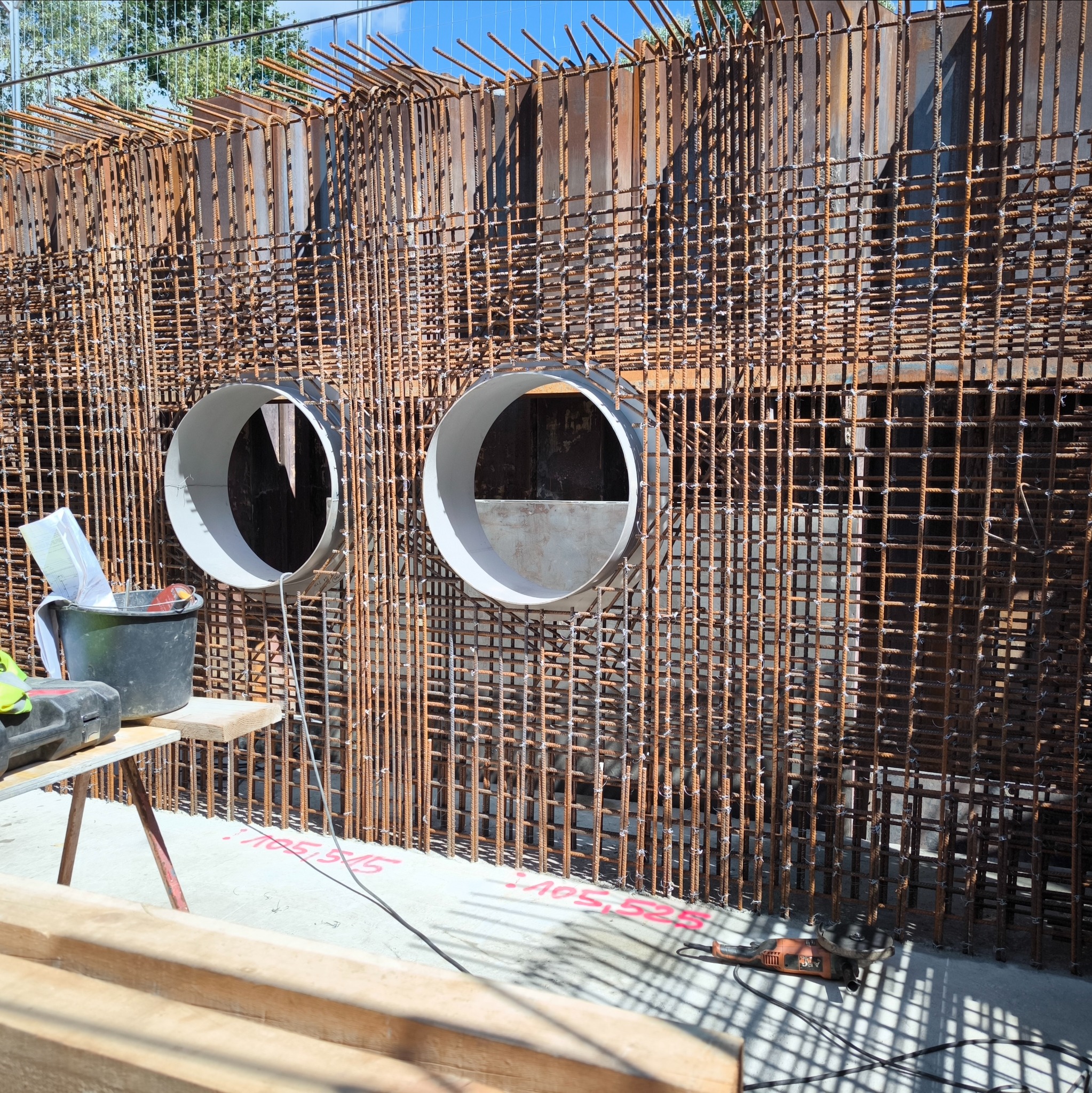 Zbrojenie ściany żelbetowej z dwoma okrągłymi otworami na przewody, widoczne narzędzia i akcesoria budowlane obok konstrukcji.