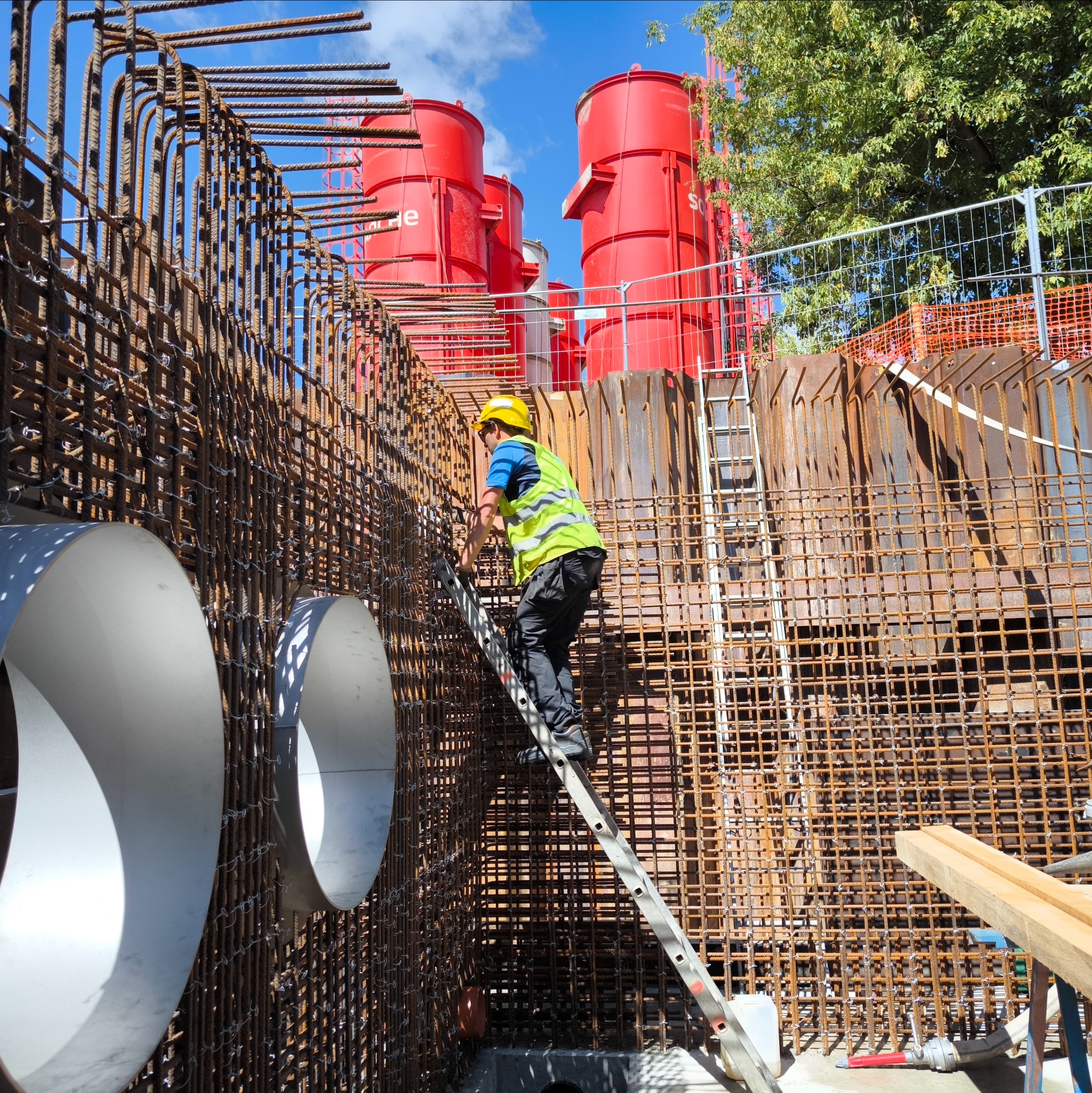 Pracownik w kasku i kamizelce na drabinie wewnątrz konstrukcji z prętów zbrojeniowych, z białymi rurami i czerwonymi silosami w tle, na placu budowy.