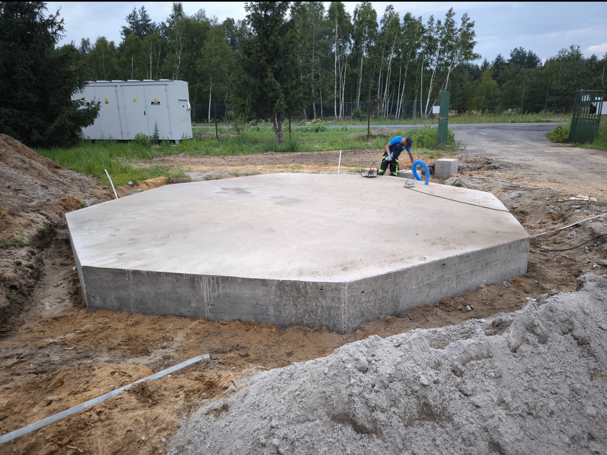 Wieloboczna płyta betonowa, prawdopodobnie pod budowę, otoczona wykopem i stertami ziemi, z pracownikiem używającym narzędzia do obróbki powierzchni, transformator w tle.