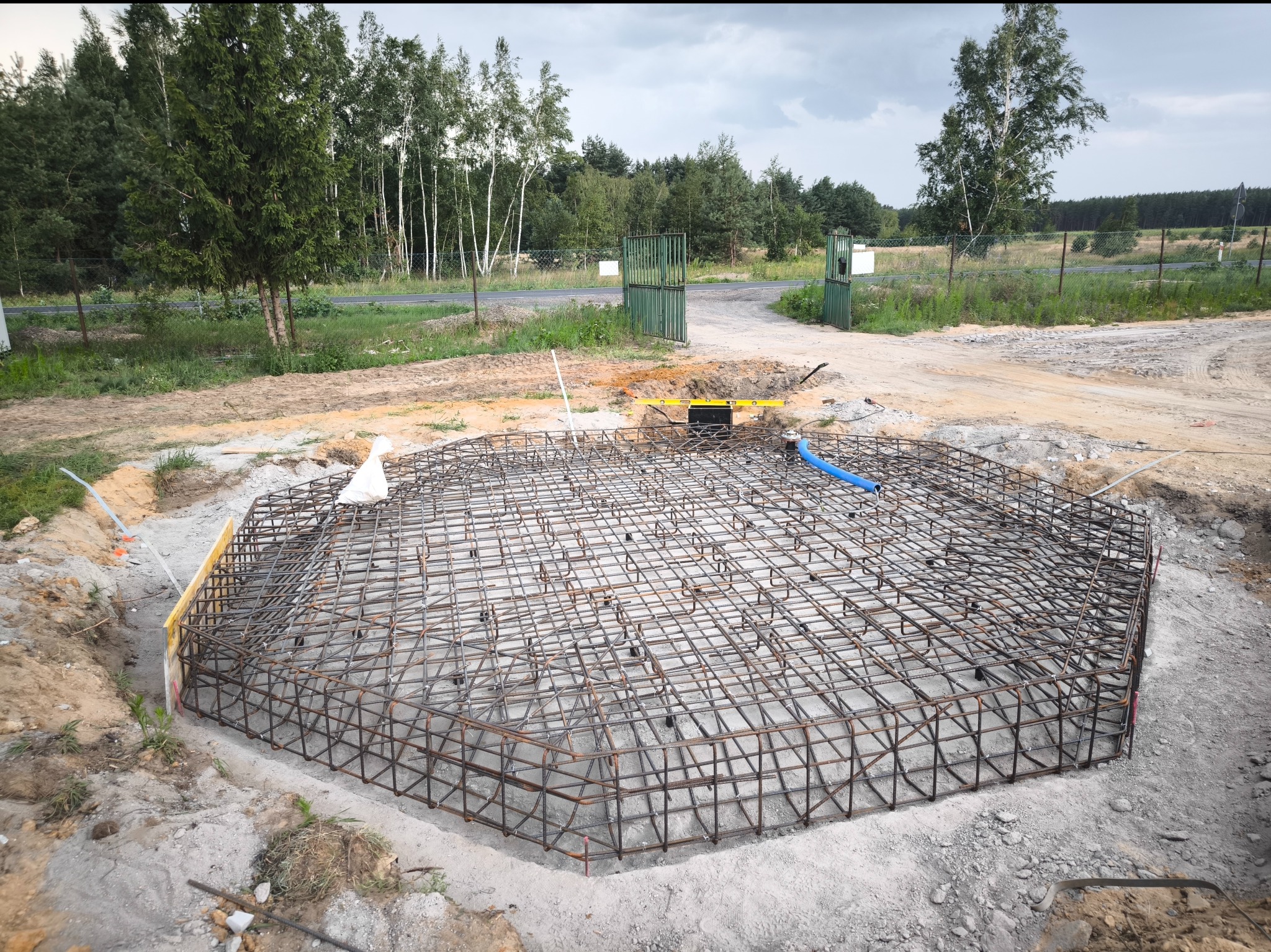 Szkielet z prętów zbrojeniowych w wykopie, przygotowany do zalania betonem, z widocznymi deskami szalunkowymi i poziomnicą w tle, na tle zieleni i pochmurnego nieba.