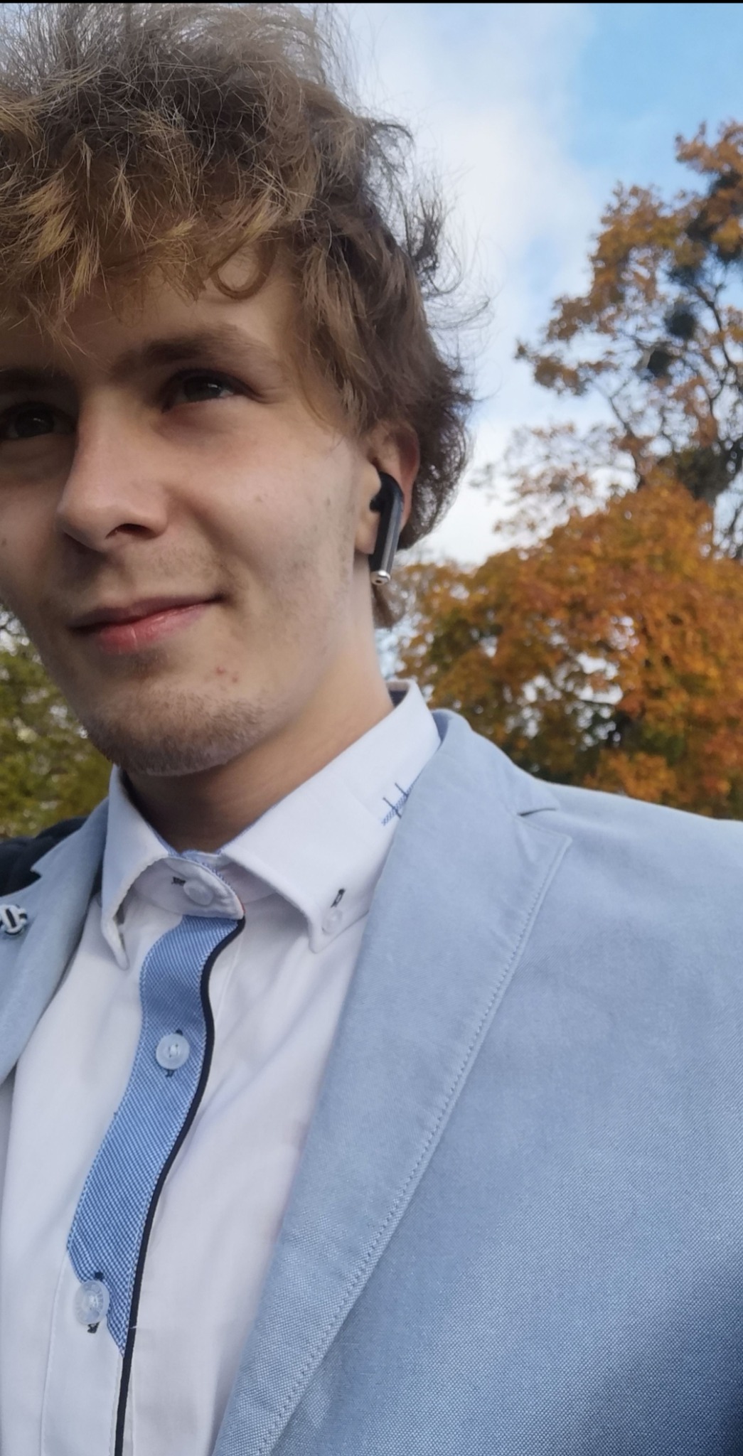 Młody mężczyzna w jasnoniebieskiej marynarce i słuchawkach bezprzewodowych, uśmiecha się na tle drzew z jesiennymi liśćmi.