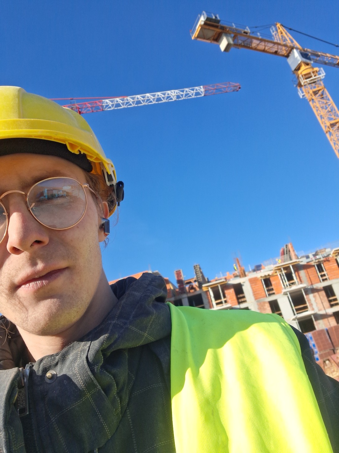 Selfie pracownika budowlanego w kasku i kamizelce odblaskowej na tle budowy z żurawiami. W okularach odbija się budynek w trakcie wznoszenia.
