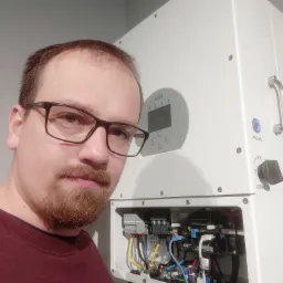 Mężczyzna w okularach pozuje obok białej skrzynki z otwartym panelem elektrycznym, widoczne kable i złącza wewnątrz.