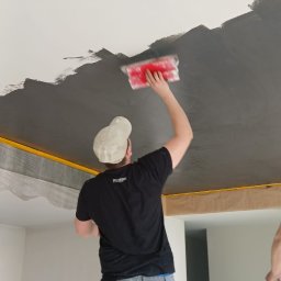 Ekipa Malarska - Pracownik w trakcie szpachlowania sufitu na ciemny kolor, widoczny z dołu, z narzędziem w ręku. Sufit częściowo pokryty, z taśmą zabezpieczającą przy ścianach.