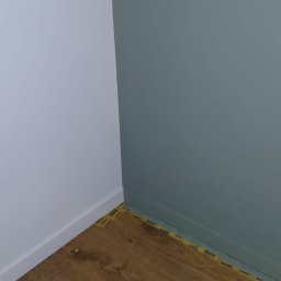 Ekipa Malarska - Świeżo pomalowane ściany w odcieniach bieli i szaro-zieleni, z nowo ułożonymi panelami podłogowymi zabezpieczonymi taśmą malarską przy listwach przypodłogowych.