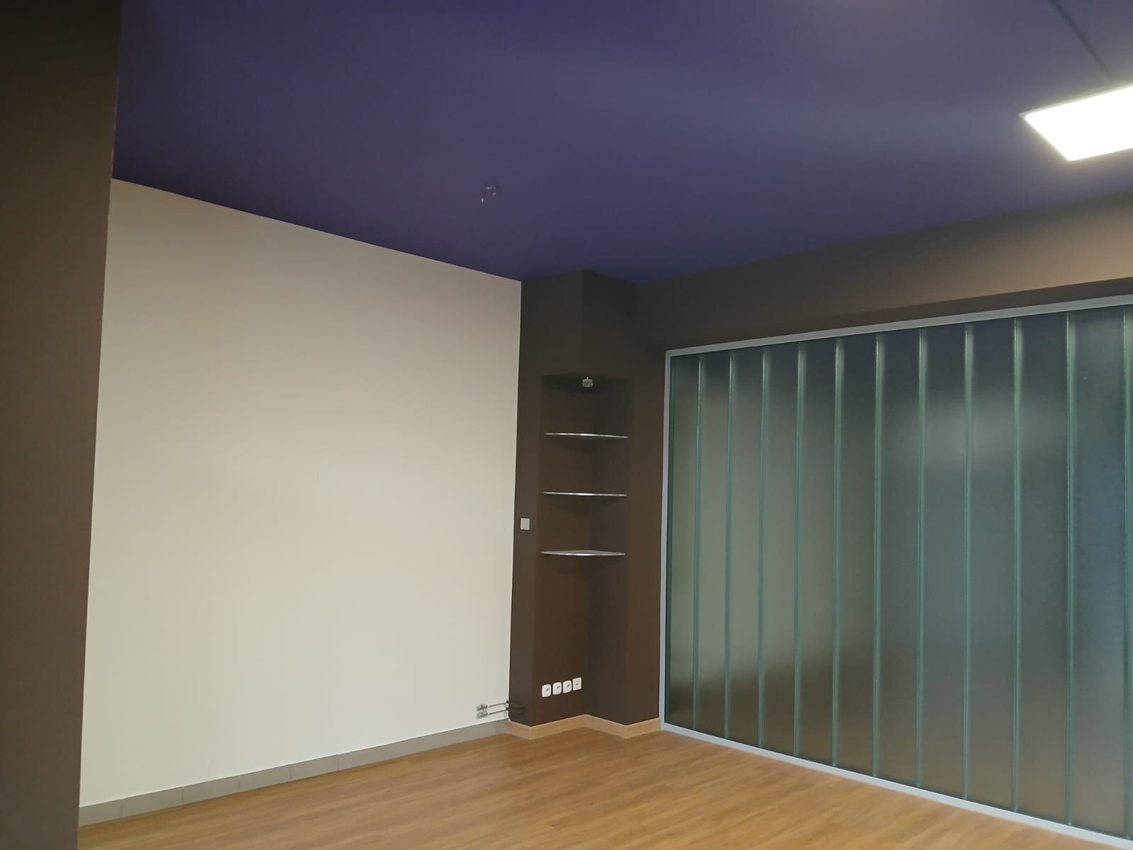 Pomieszczenie po remoncie: biała ściana, fioletowy sufit, brązowe elementy, szklana ściana działowa z pionowymi panelami i drewniana podłoga. Widoczne półki w rogu.