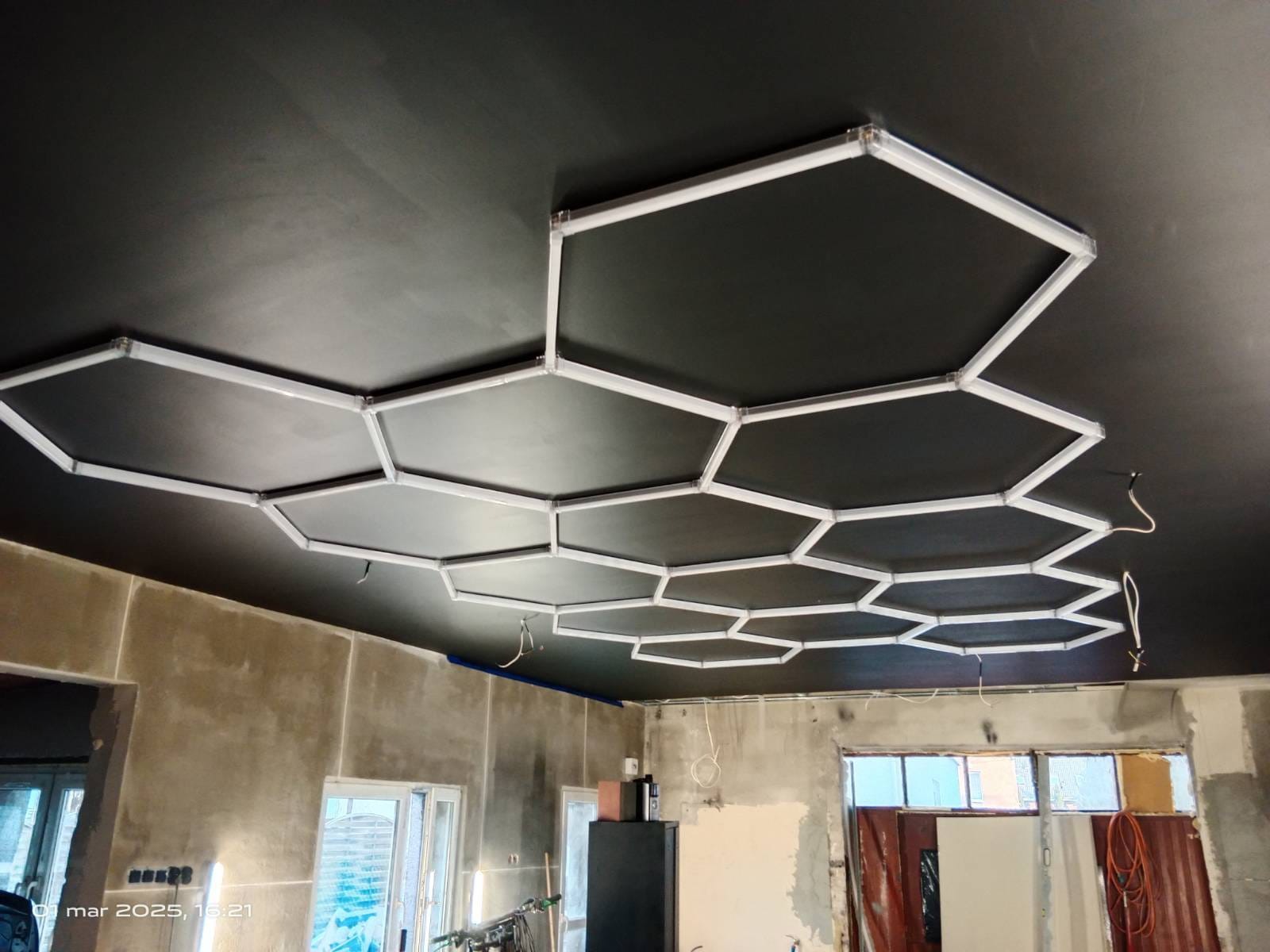 Czarny sufit z heksagonalnymi panelami LED w surowym wnętrzu. Widoczne kable i niedokończone ściany, co sugeruje etap prac wykończeniowych.