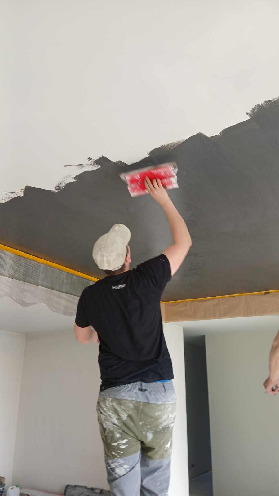 Pracownik w trakcie szpachlowania sufitu na ciemny kolor, widoczny z dołu, z narzędziem w ręku. Sufit częściowo pokryty, z taśmą zabezpieczającą przy ścianach.