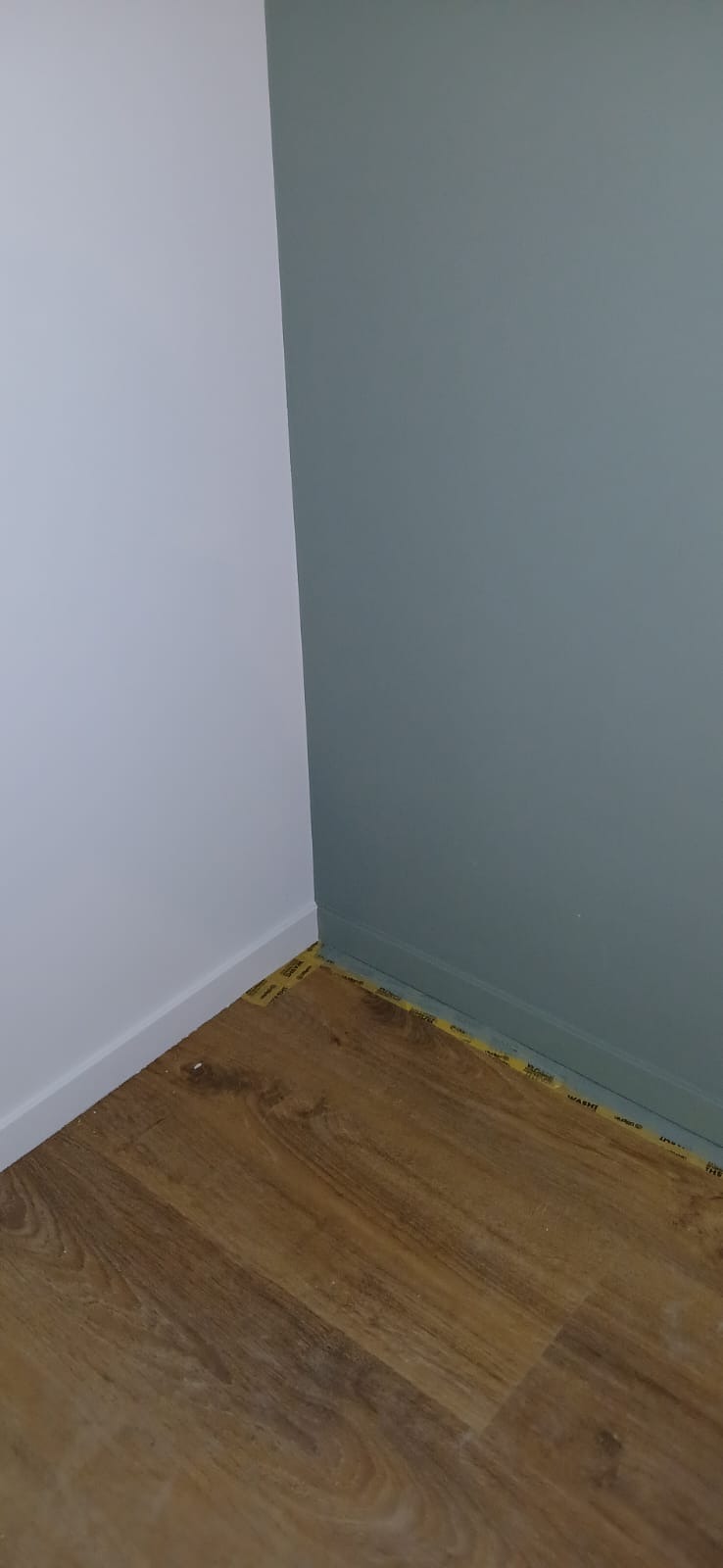 Świeżo pomalowane ściany w odcieniach bieli i szaro-zieleni, z nowo ułożonymi panelami podłogowymi zabezpieczonymi taśmą malarską przy listwach przypodłogowych.