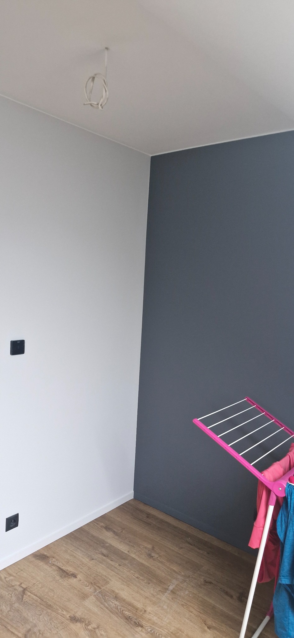 Świeżo pomalowane wnętrze z szarą ścianą, drewnianą podłogą i suszarką na ubrania. Widoczne gniazdka elektryczne i wystające kable od oświetlenia.