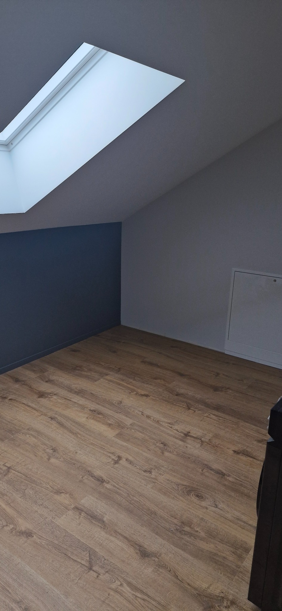 Pokój na poddaszu z drewnianą podłogą, pomalowaną ścianą i oknem dachowym. Widoczny fragment mebla w prawym dolnym rogu. Ujęcie z niskiego kąta.