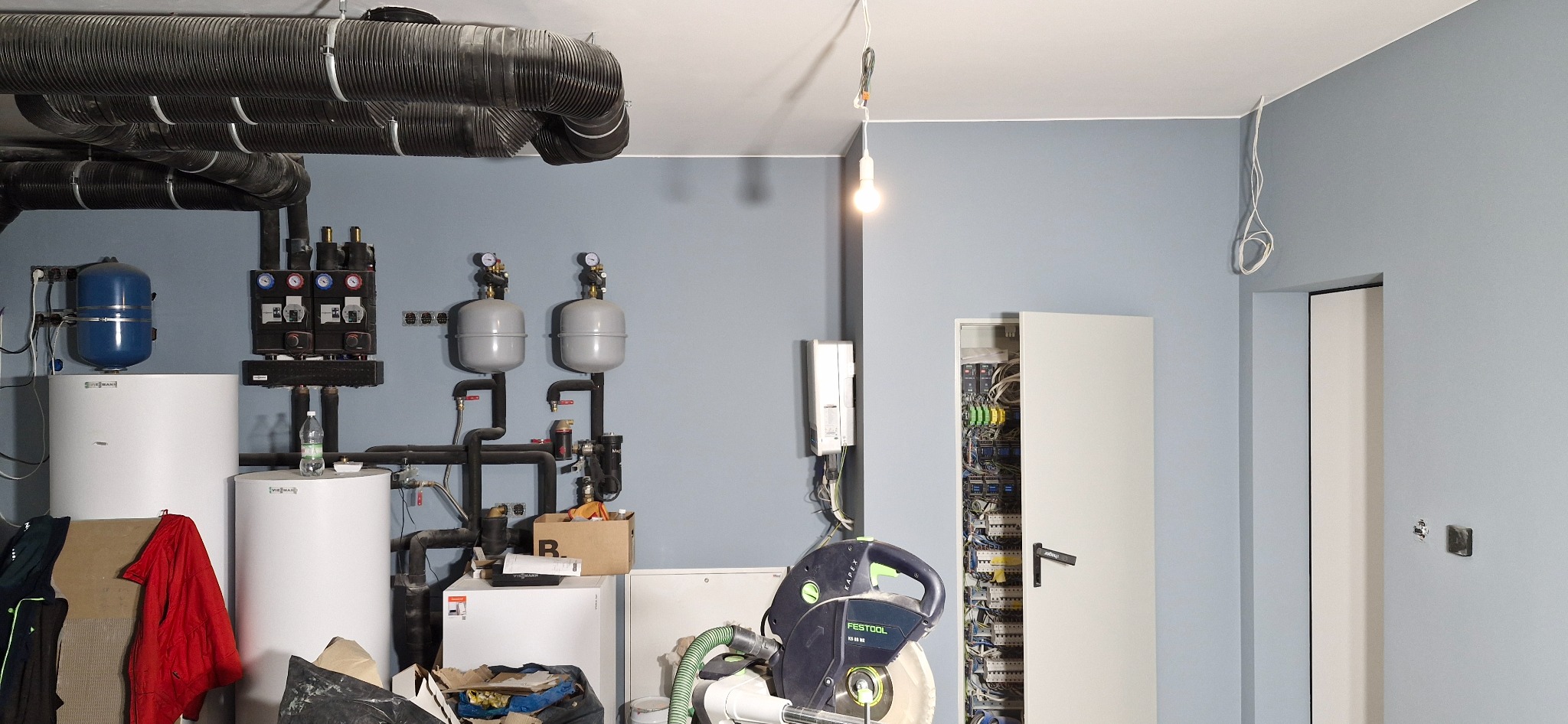Kotłownia w trakcie prac: widoczne bojlery, rury, rozdzielnice, narzędzia (piła Festool). Ściany pomalowane na niebiesko, sufit biały z widocznymi rurami wentylacyjnymi.