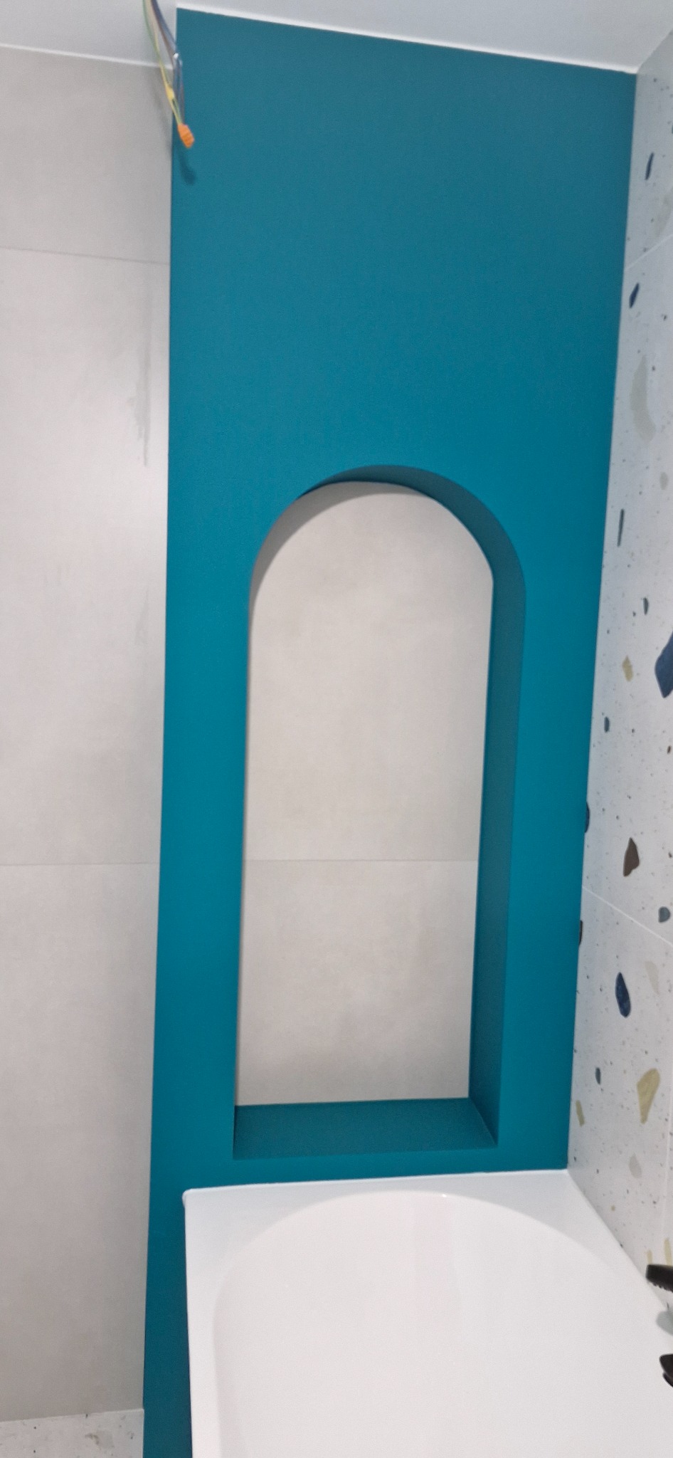 Niebieska wnęka z zaokrąglonym łukiem w wykończonej łazience, nad białą wanną. Widoczne płytki w dwóch wzorach i wystające kable.