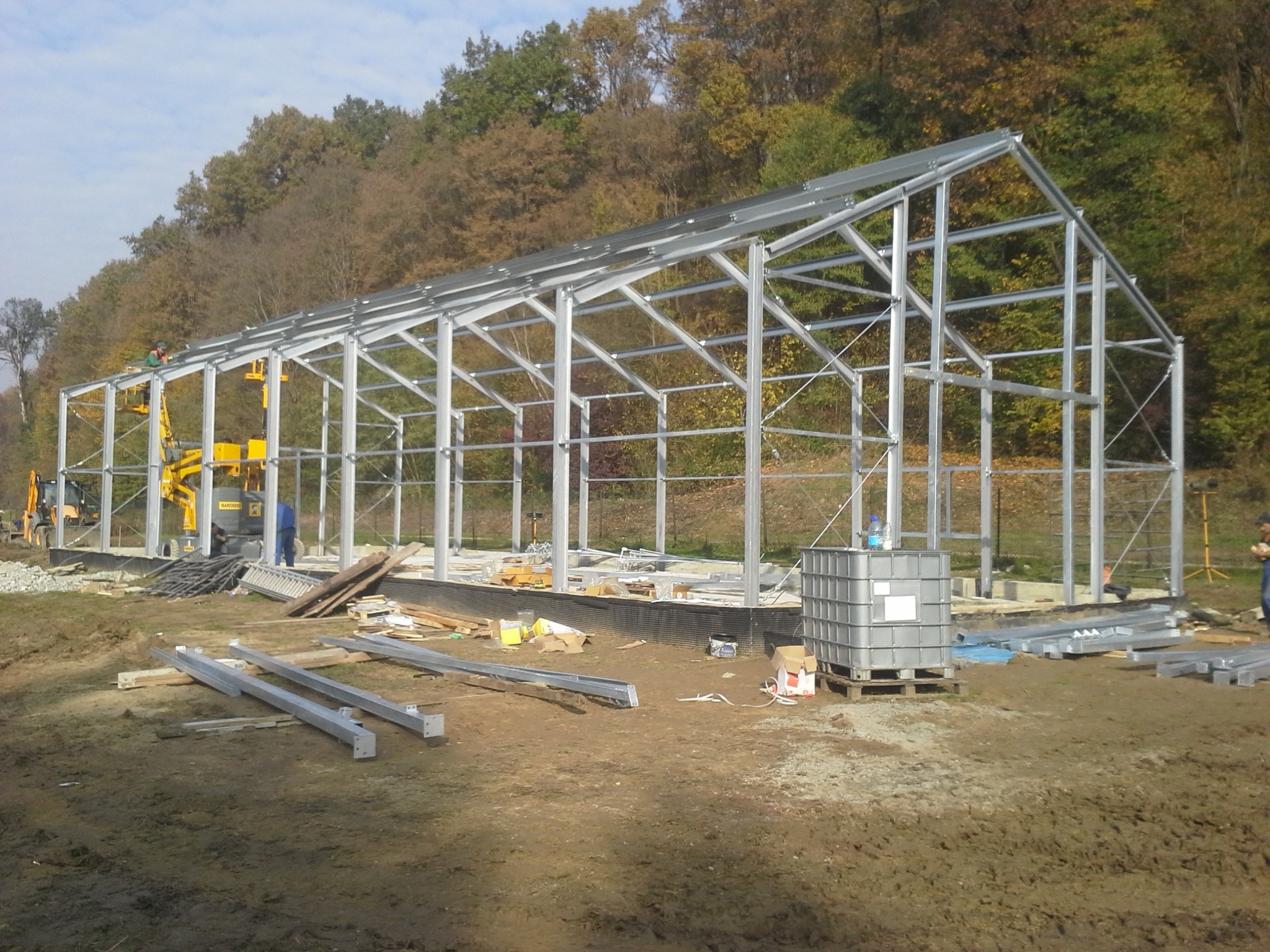 Montaż szkieletu hali stalowej na fundamencie, widoczne elementy konstrukcyjne, podnośnik koszowy i pracownicy w tle drzew.