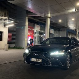 DK-company - Czarny Lexus ES z numerem rejestracyjnym ZS 737MC stoi przed terminalem lotniska nocą, z widocznym wejściem i taksówką w tle.