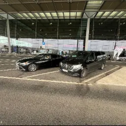 Dwa czarne samochody, sedan Lexus i van Mercedes, zaparkowane przed nowoczesnym budynkiem lotniska z dużymi szklanymi fasadami i napisem 'FREIHEIT IST' widocznym w tle.