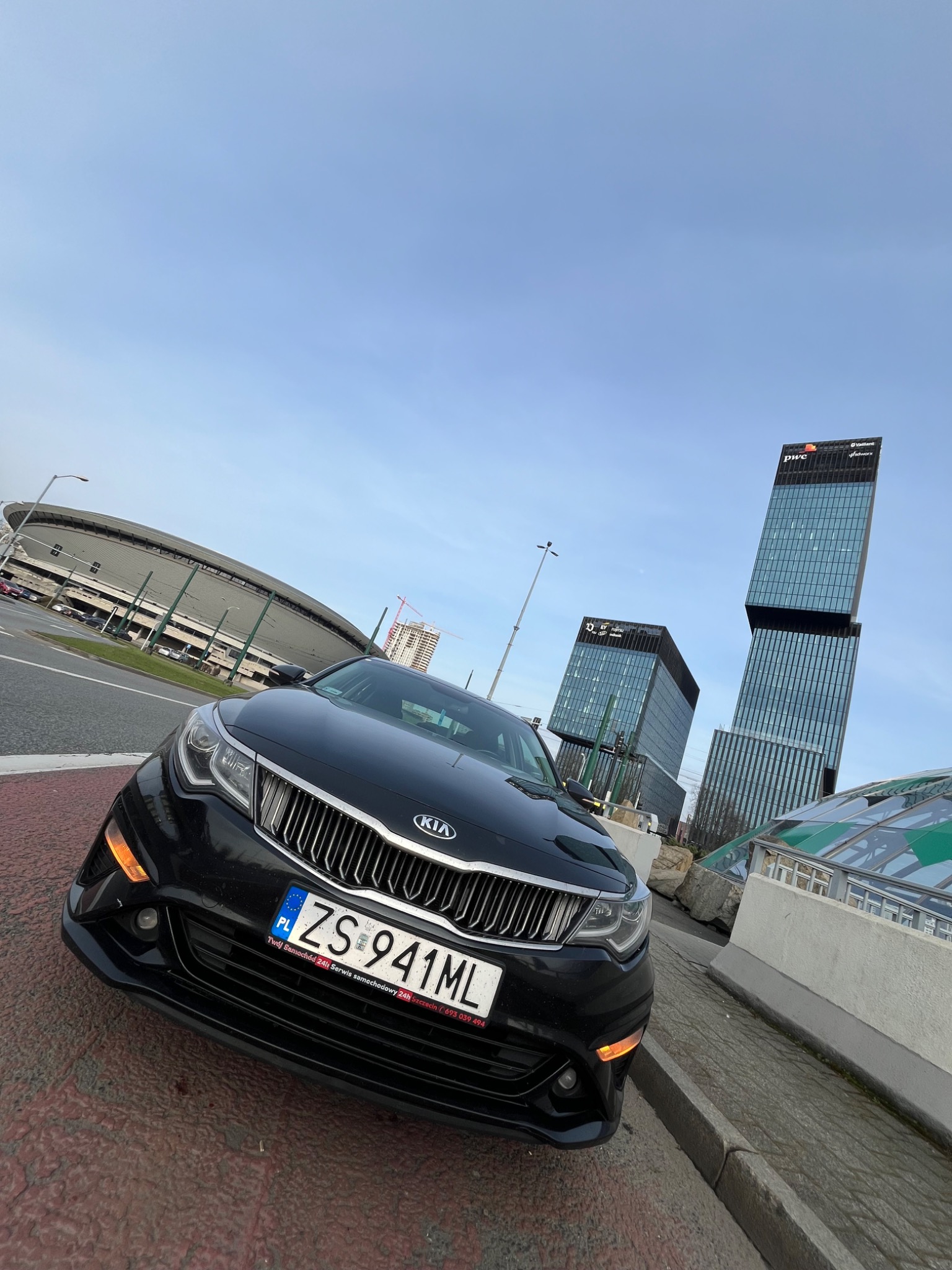 Czarny samochód Kia z polską tablicą rejestracyjną ZS 941ML na tle stadionu i nowoczesnych biurowców w Szczecinie.