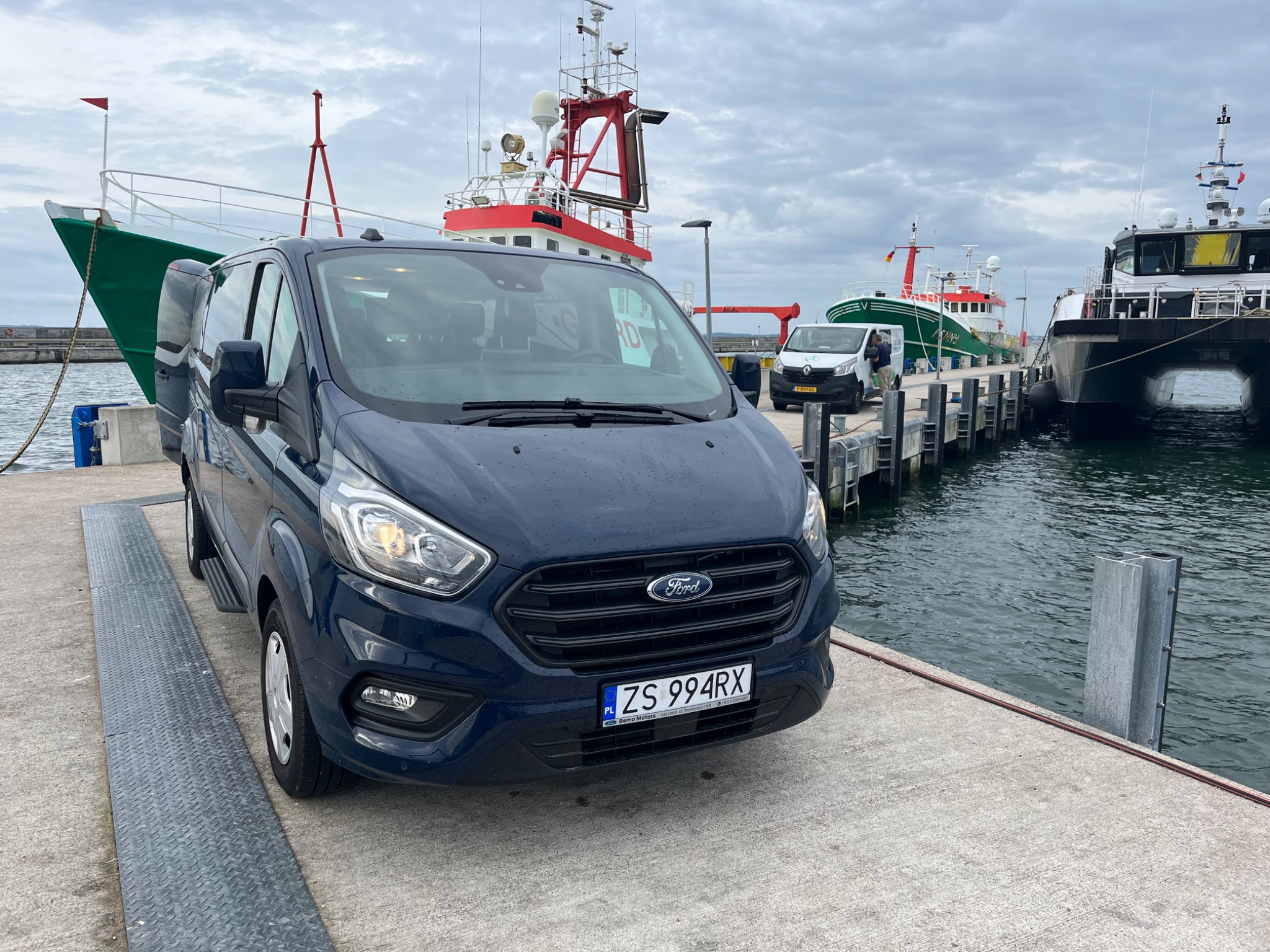 Niebieski van Ford Transit Custom zaparkowany na nabrzeżu portowym, z widocznymi statkami rybackimi w tle, tablica rejestracyjna ZS 994RX.