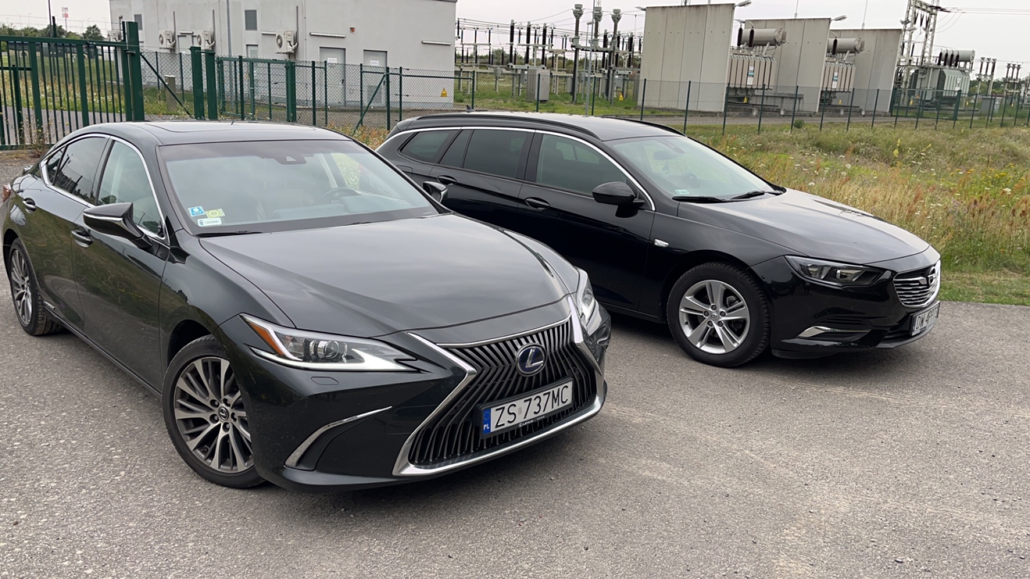 Dwa czarne samochody: Lexus ES z numerem rejestracyjnym ZS 737MC i Opel Insignia kombi, zaparkowane na szarej nawierzchni z budynkami transformatorowymi w tle.