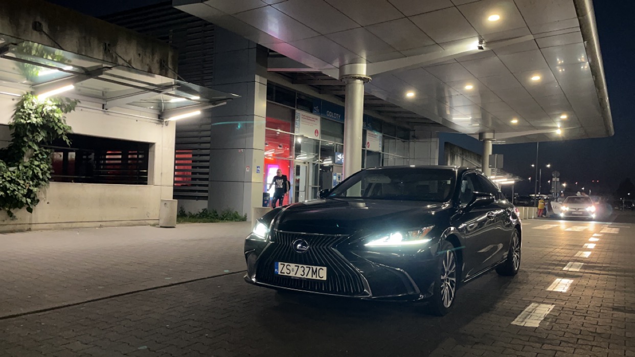 Czarny Lexus ES z numerem rejestracyjnym ZS 737MC stoi przed terminalem lotniska nocą, z widocznym wejściem i taksówką w tle.