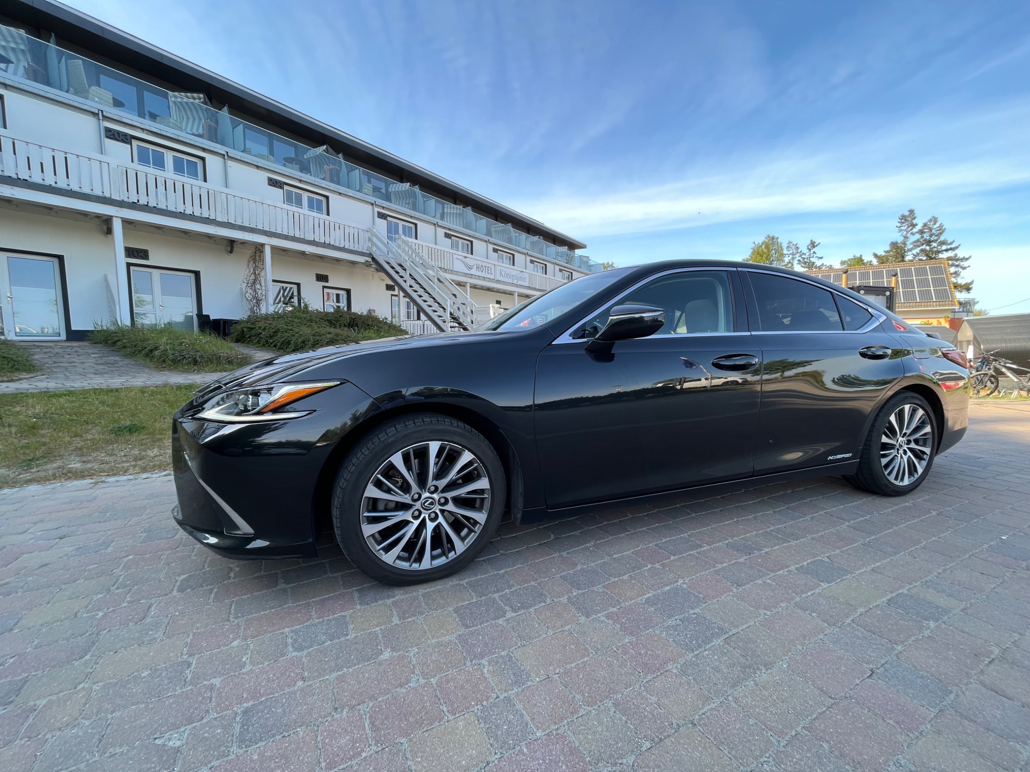 Czarny sedan Lexus ES 300h zaparkowany przed hotelem z balkonami i schodami zewnętrznymi, widok z boku na kostce brukowej.