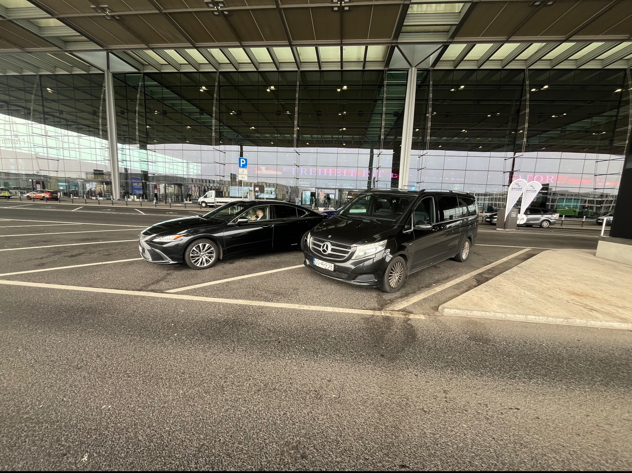 Dwa czarne samochody, sedan Lexus i van Mercedes, zaparkowane przed nowoczesnym budynkiem lotniska z dużymi szklanymi fasadami i napisem 'FREIHEIT IST' widocznym w tle.