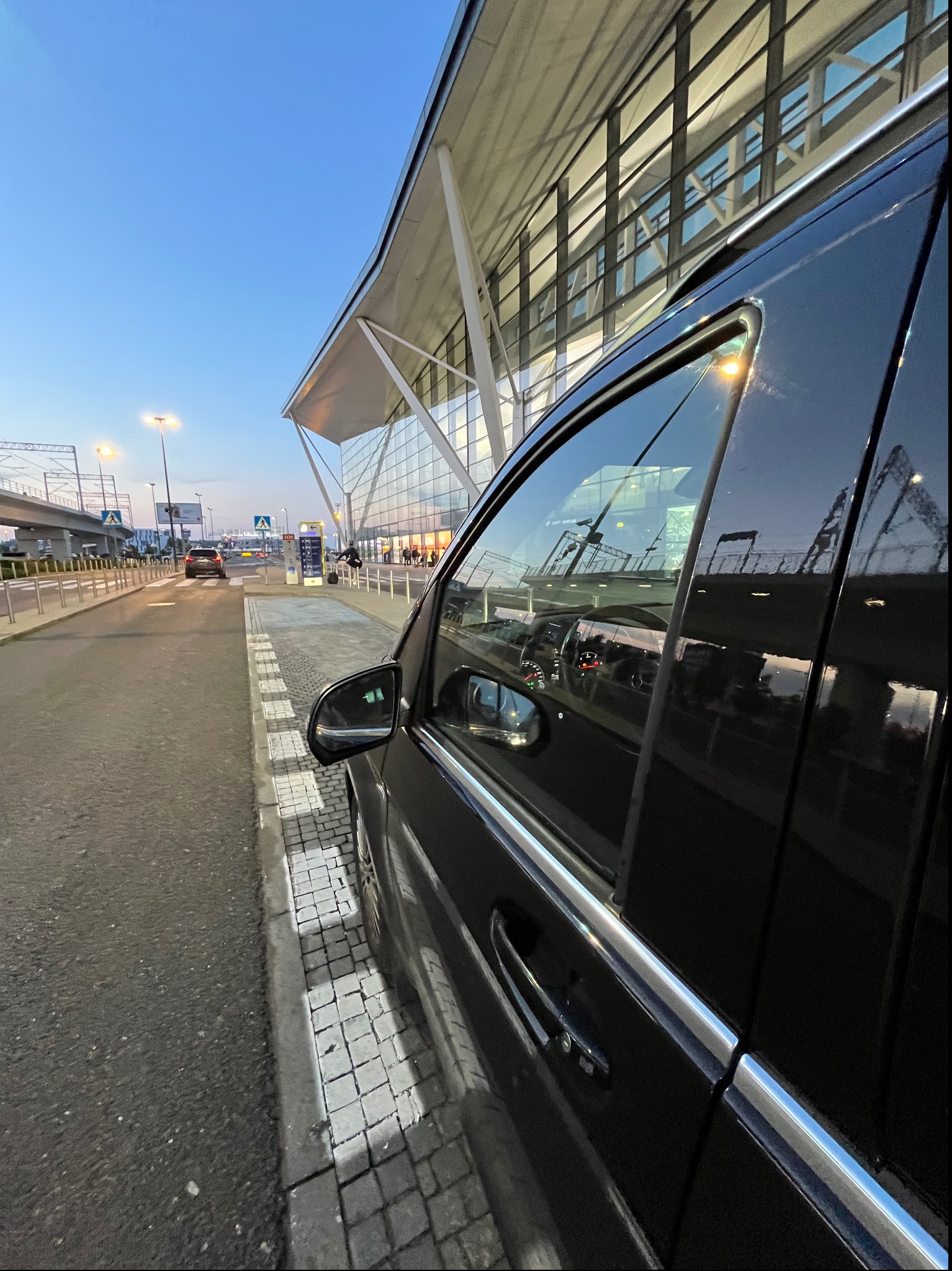 Czarna limuzyna zaparkowana przed nowoczesnym terminalem lotniska o zmierzchu, widoczny fragment wnętrza i odbicie otoczenia w szybach.