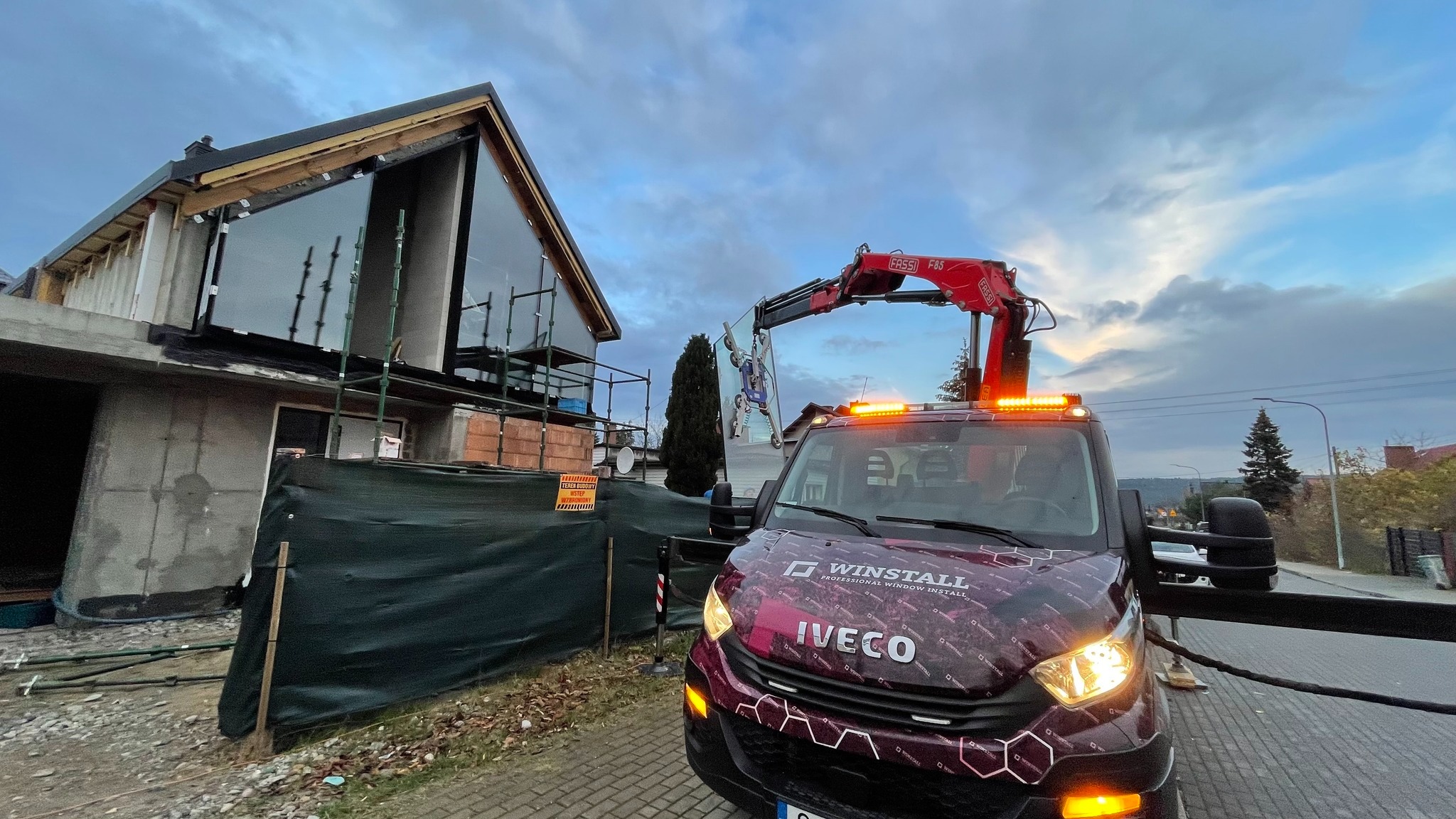 Montaż dużej szyby na budowie domu jednorodzinnego przy użyciu specjalistycznego dźwigu samochodowego z przyssawkami; widoczny budynek w stanie surowym otwartym, rusztowania i samochód Iveco z logo...