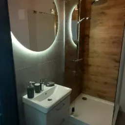 Nowoczesna łazienka z kabiną prysznicową typu walk-in, umywalką z szafką i okrągłym lustrem podświetlanym LED, wykończona płytkami imitującymi drewno i szarym gresem.