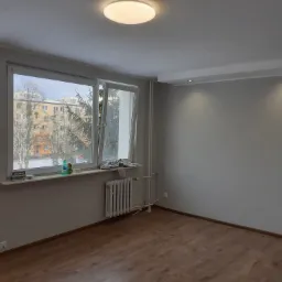 Pomalowane na szaro wnętrze pokoju z drewnianą podłogą, widoczne okno z widokiem na budynki i drzewa, grzejnik pod oknem, gładkie ściany i sufit z oświetleniem punktowym.