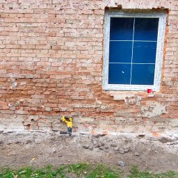 Marcin kwasnik usługi ogolnobudowlane - Dwa okna PCV z białymi ramami w surowej, ceglanej ścianie, widoczne ślady napraw i narzędzia, w tym młot pneumatyczny i poziomica.