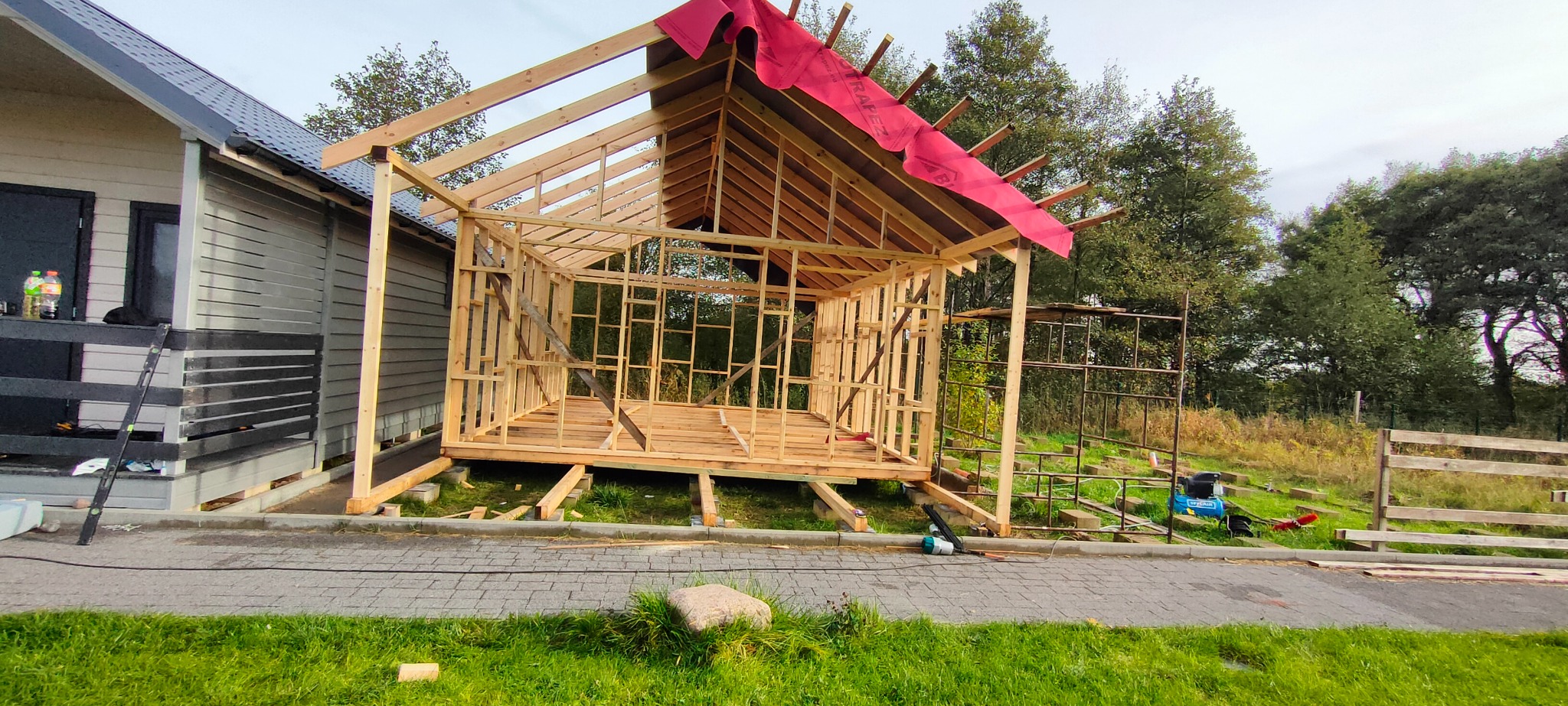Konstrukcja szkieletowa drewnianej altany lub domku letniskowego w trakcie budowy, widoczne elementy więźby dachowej i ściany szkieletowe, zabezpieczone czerwoną folią dachową.