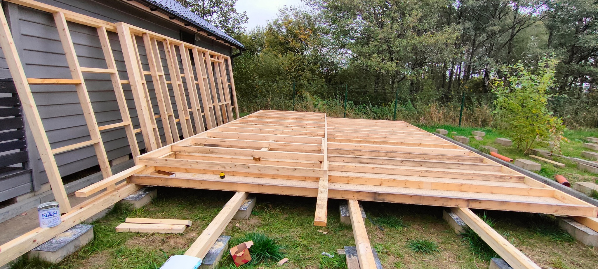 Konstrukcja drewnianej podłogi i ściany szkieletowej w trakcie budowy na zewnątrz, widoczne betonowe bloczki fundamentowe, puszka farby i narzędzia.