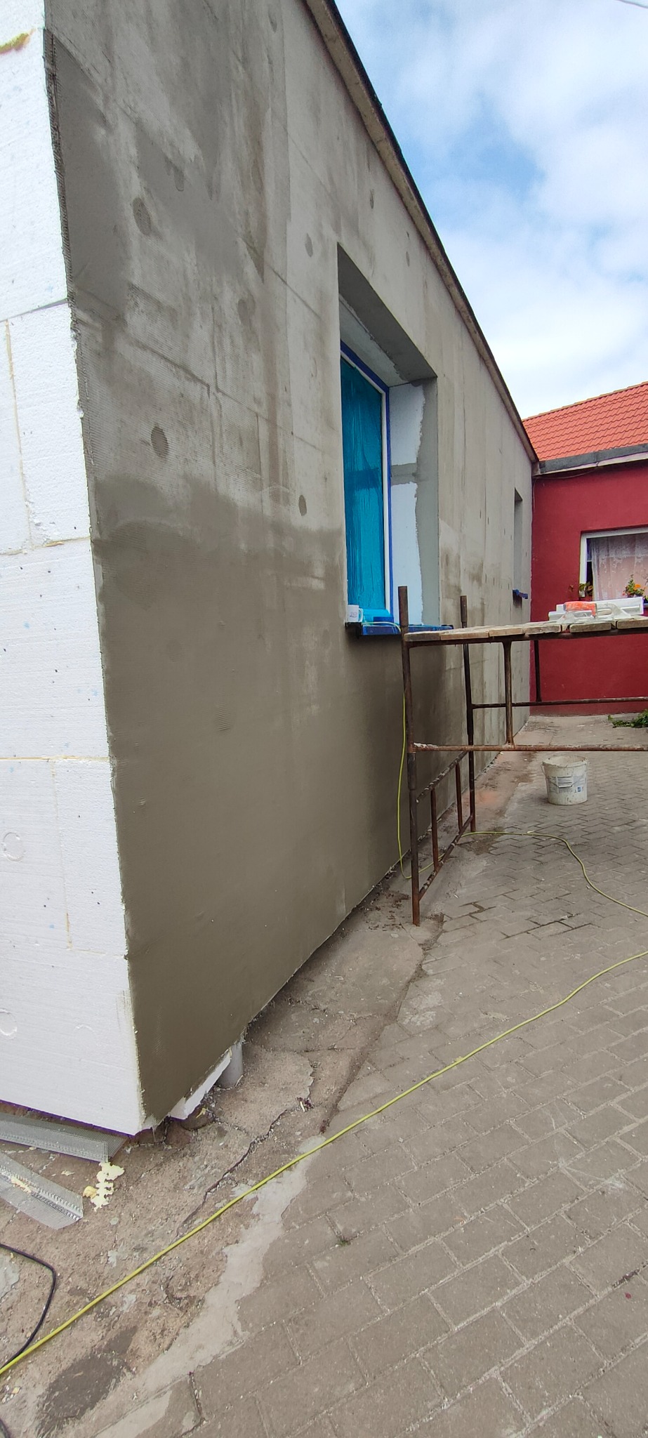 Ocieplanie budynku w trakcie realizacji: fragment ściany z ociepleniem styropianowym i nałożoną warstwą szarej zaprawy, okno zabezpieczone folią, widoczne rusztowanie i wiadro z zaprawą.