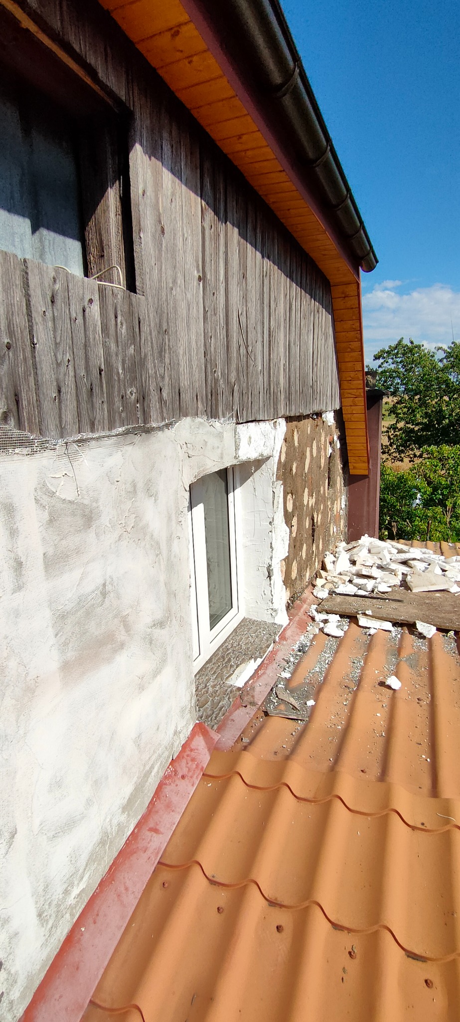 Fragment elewacji domu w trakcie ocieplania, widoczne warstwy izolacji, siatka i częściowo nałożony tynk przy oknie, z fragmentami starej elewacji z drewnianych desek powyżej.