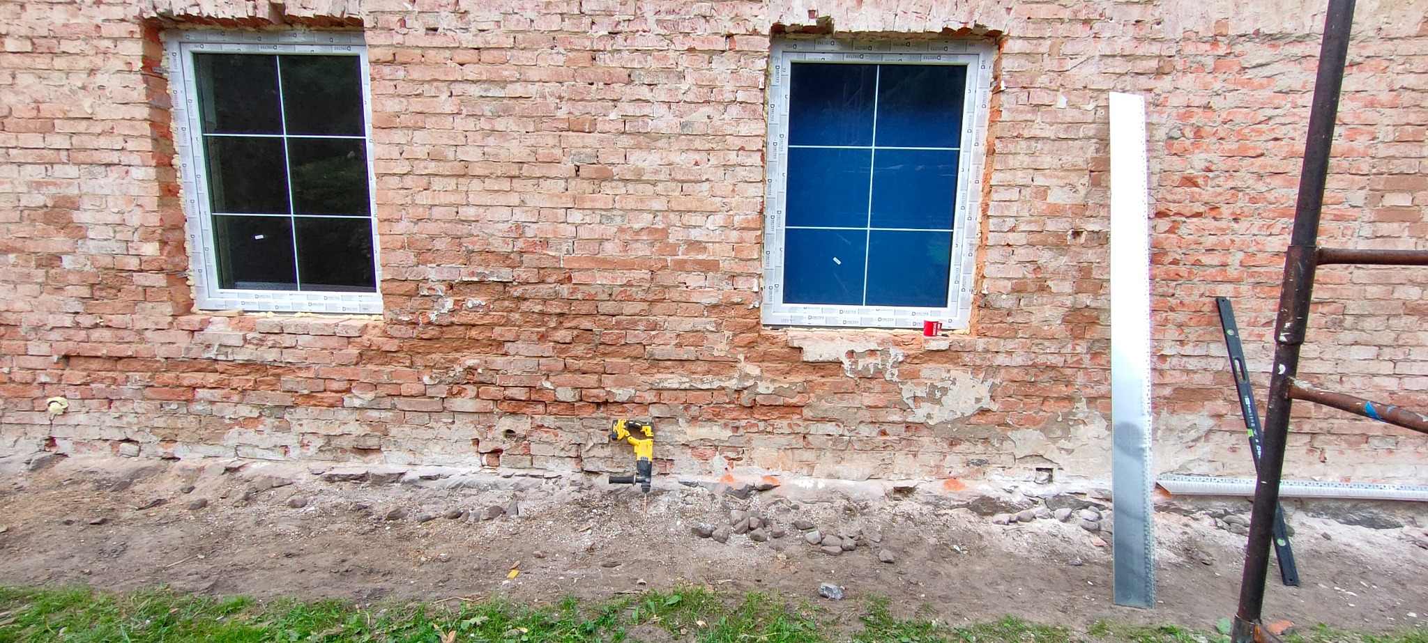 Dwa okna PCV z białymi ramami w surowej, ceglanej ścianie, widoczne ślady napraw i narzędzia, w tym młot pneumatyczny i poziomica.