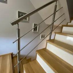 Schody drewniane z białą wstawką i nowoczesną metalową poręczą, na ścianie wiszą trzy obrazy w białych ramkach.