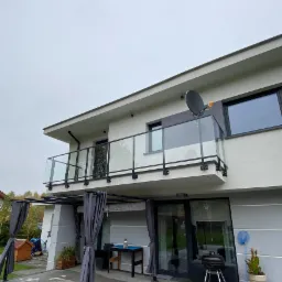 Dwupiętrowy dom z balkonem z balustradą szklaną z czarnymi elementami mocującymi, widok z dołu na szarą elewację i satelitę na dachu.